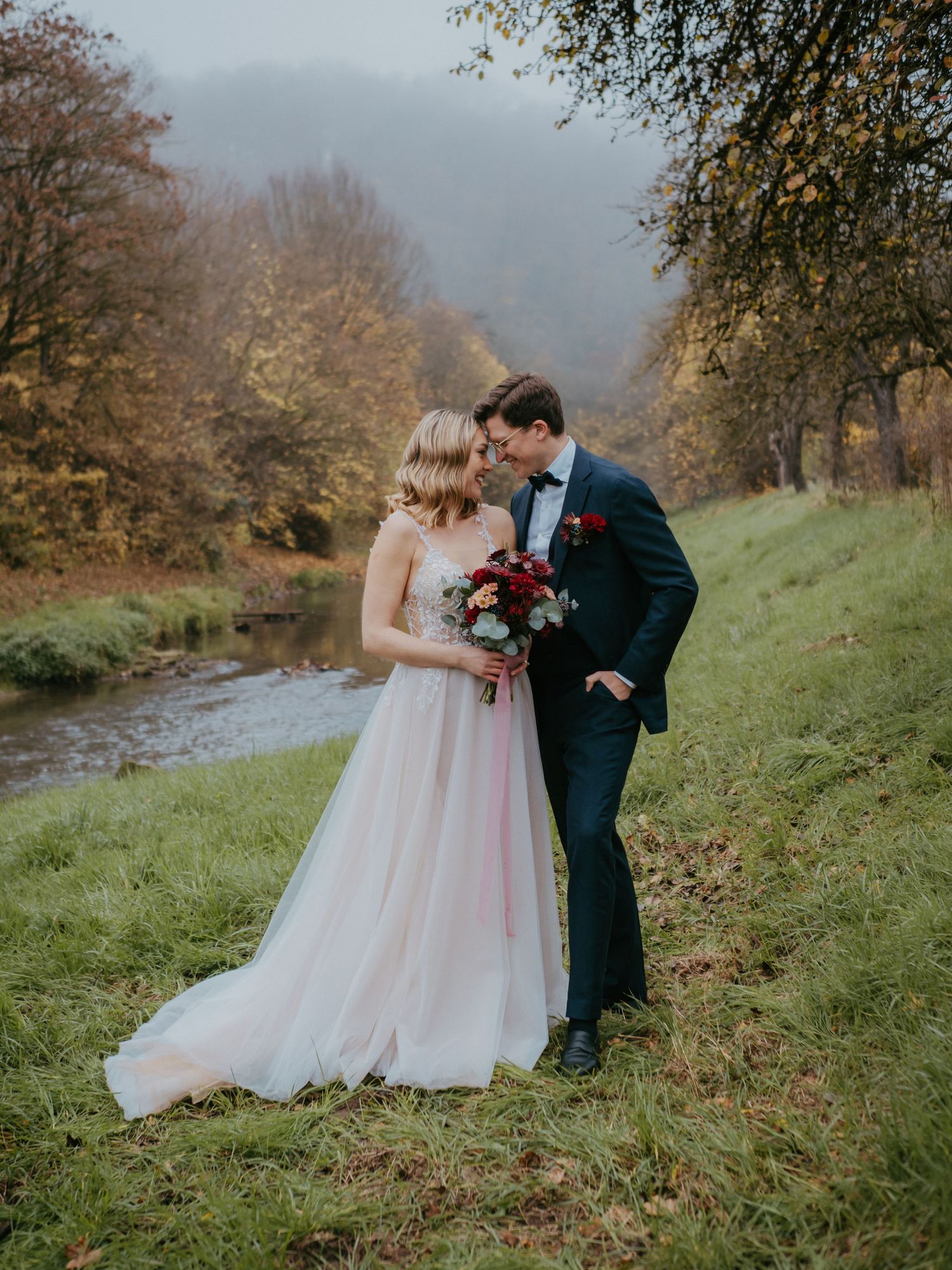 A couple shares an intimate moment on their wedding day by a stream in an autumn forest setting.