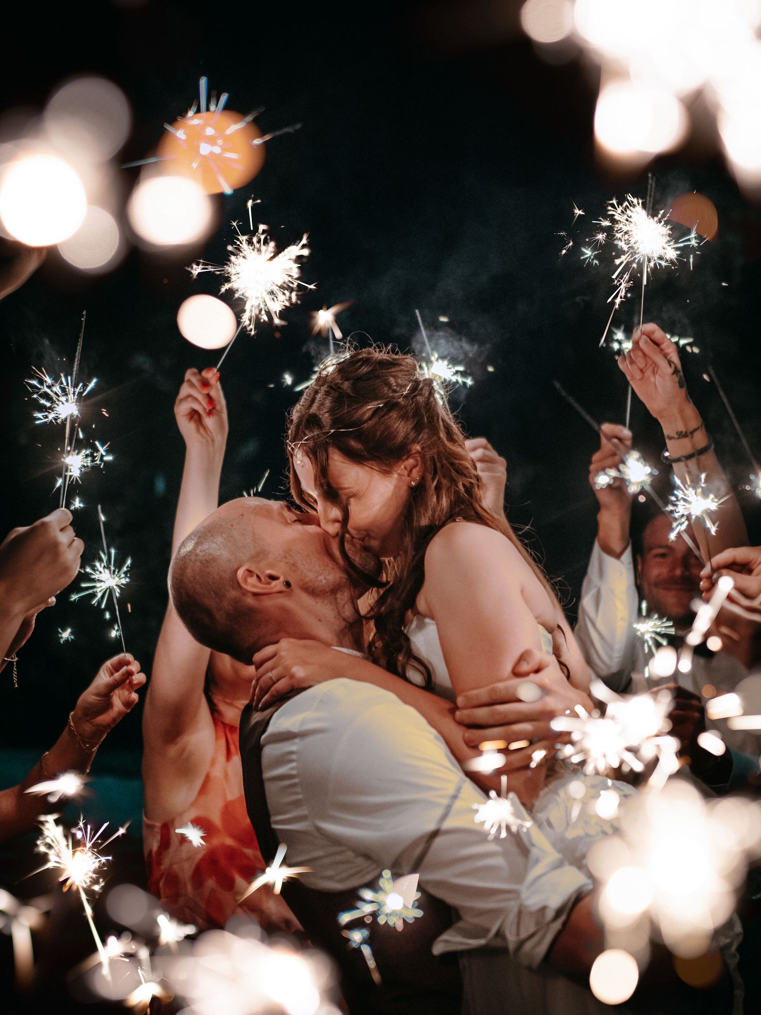 Romantic couple kissing and holding sparklers at night with glowing bokeh lights creating a magical atmosphere.