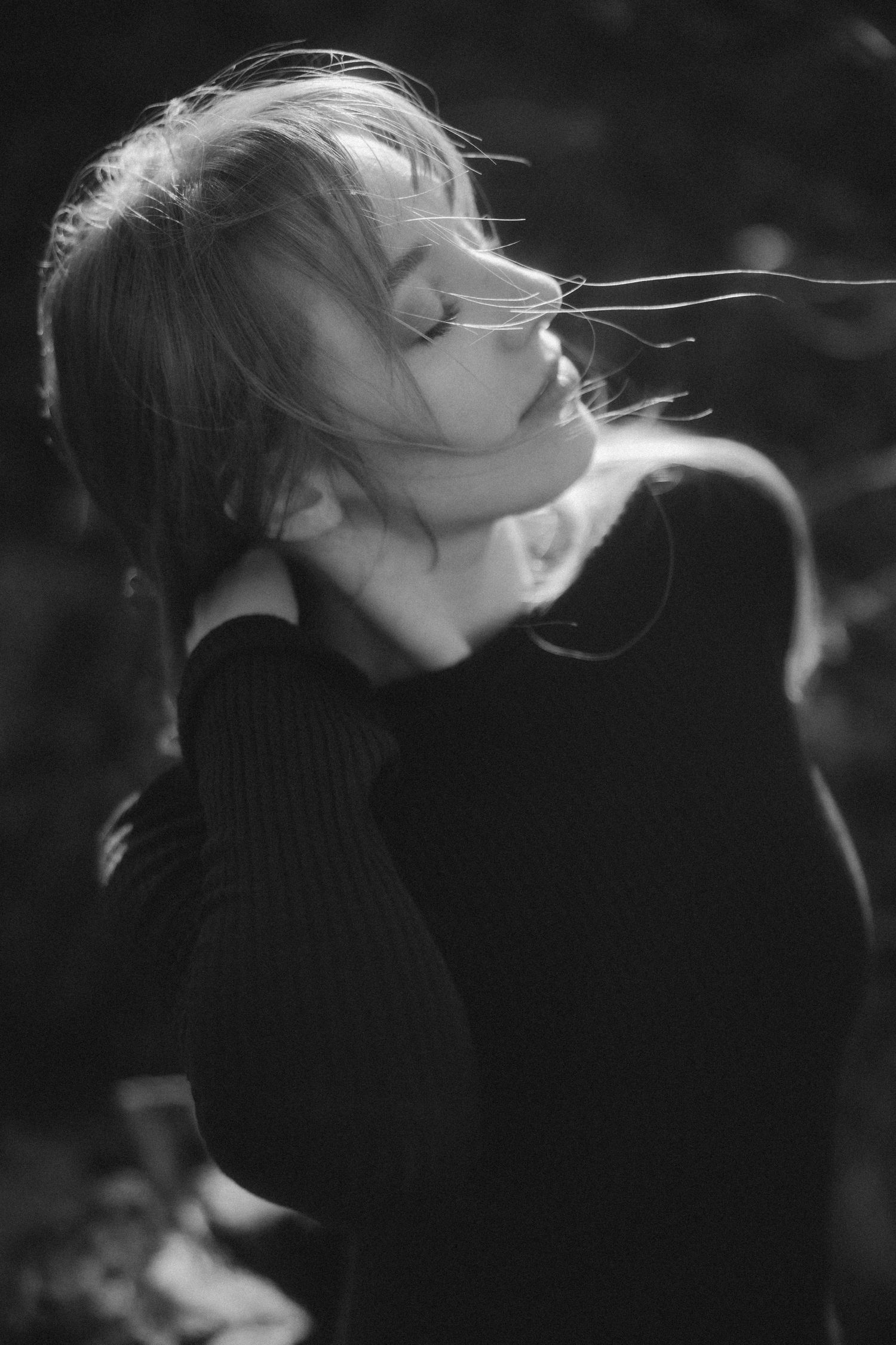 Artistic black and white portrait of a woman in a turtleneck sweater with wind-tousled hair against dark background.