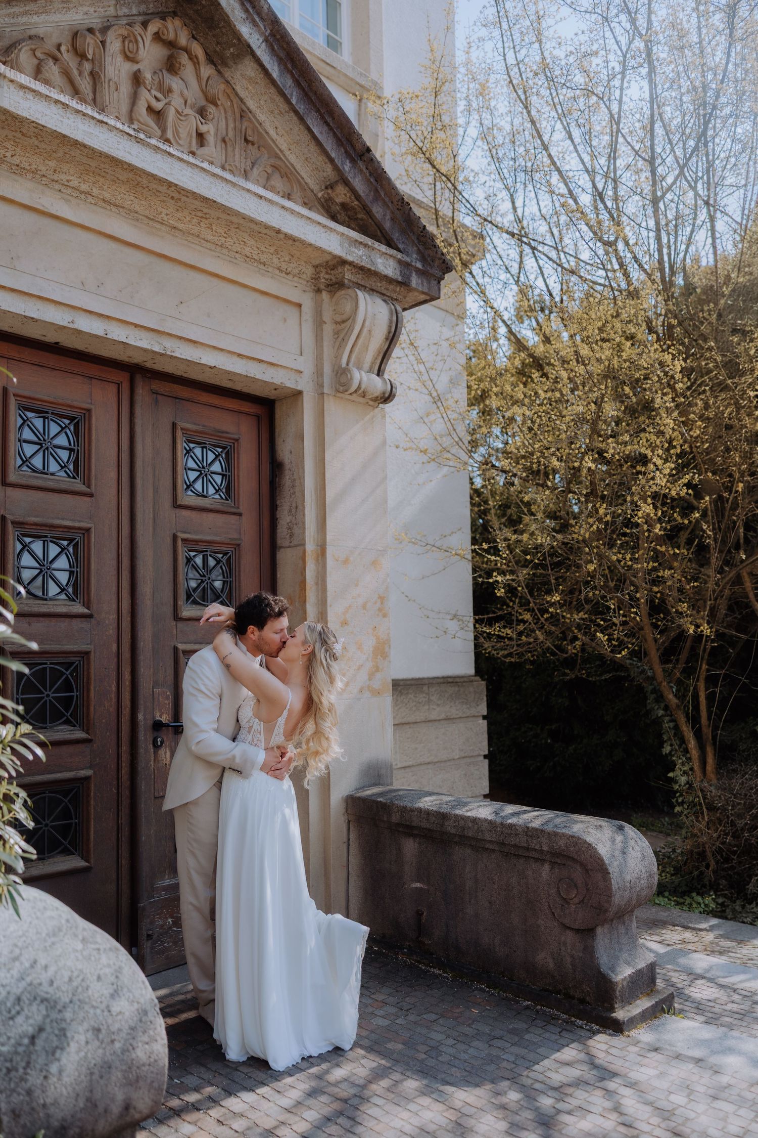 A couple in elegant white wedding attire embracing by an ornate wooden church door with decorative stone architecture.