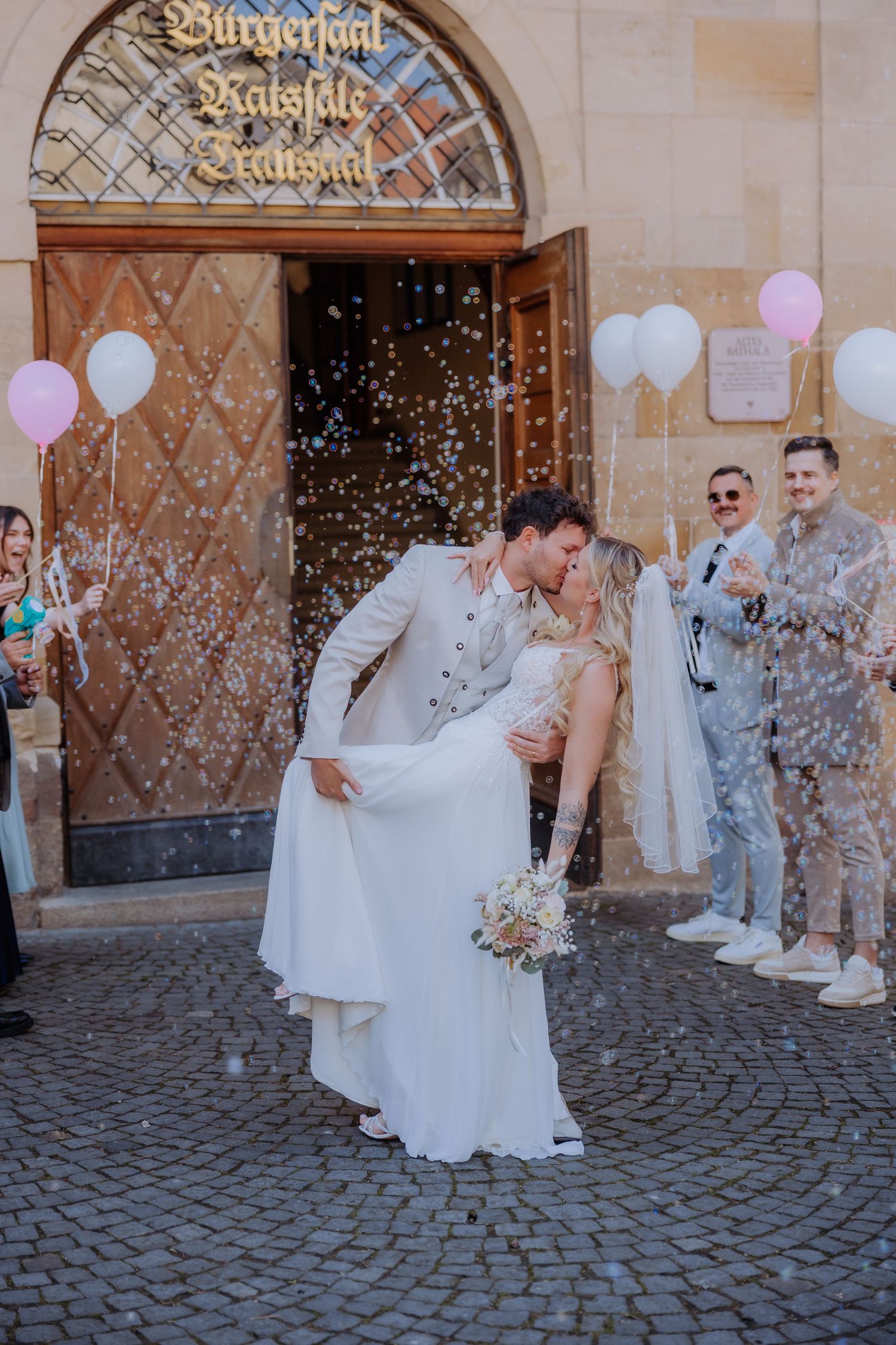 Bride and groom share romantic dip kiss outside wedding hall with confetti and pink balloons while guests celebrate.