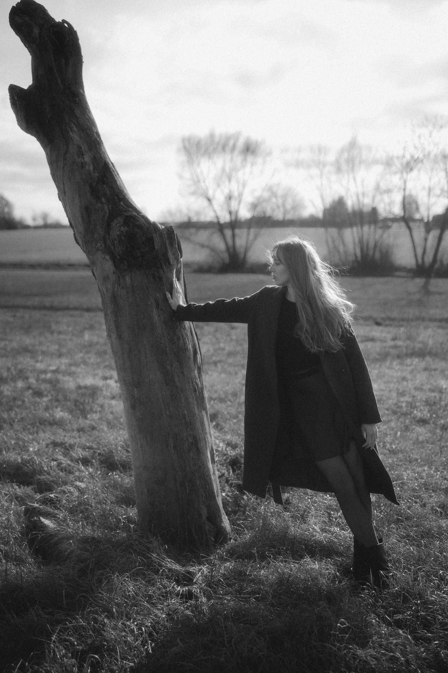 A moody black and white sequence showing a figure in dark clothing moving behind a stark tree trunk in a field.