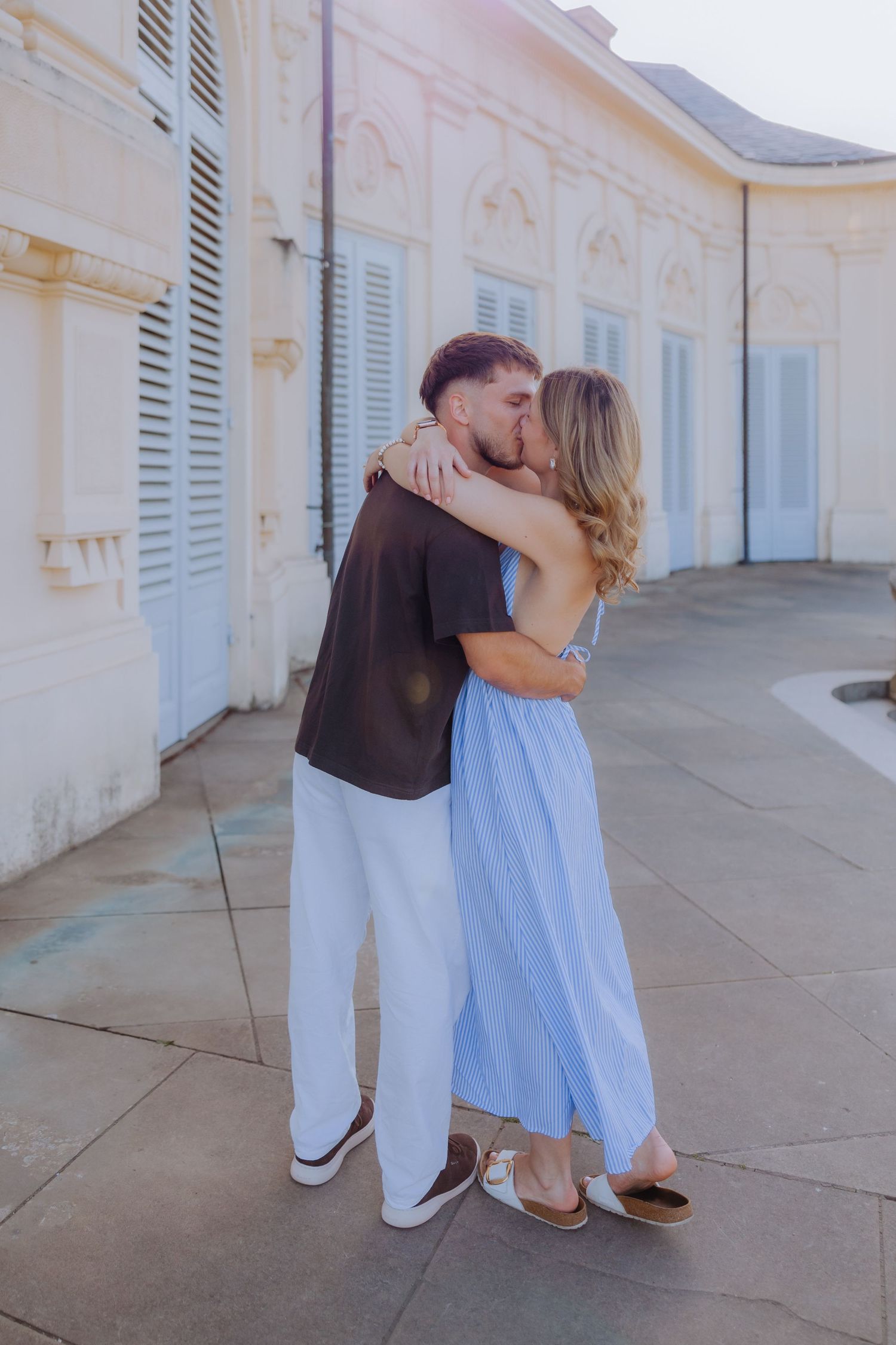 A couple shares a romantic kiss outside a elegant white building, the woman in a blue dress and man in casual attire.
