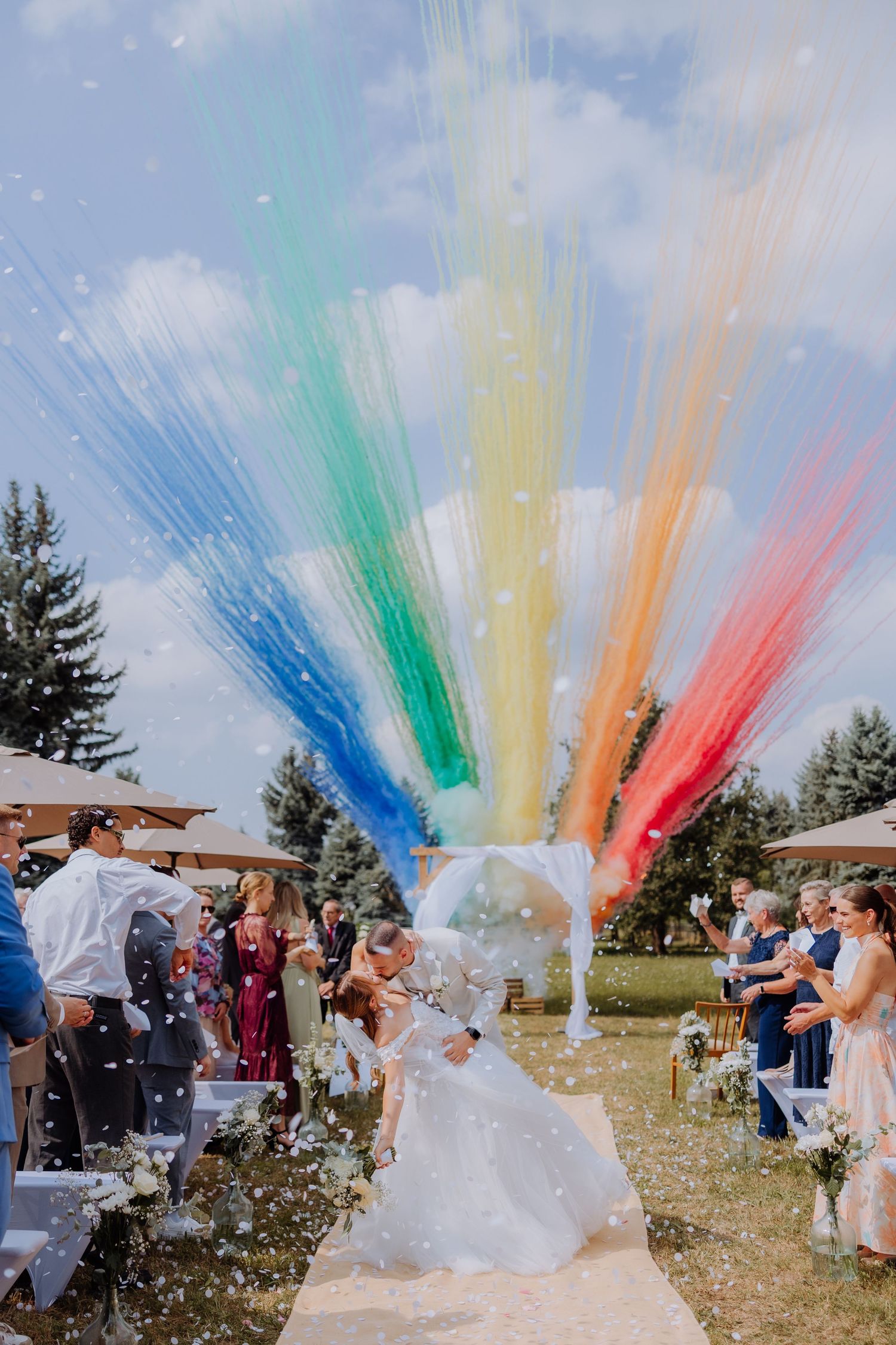 Bride and groom kiss at outdoor wedding ceremony with colorful rainbow streamers bursting overhead.