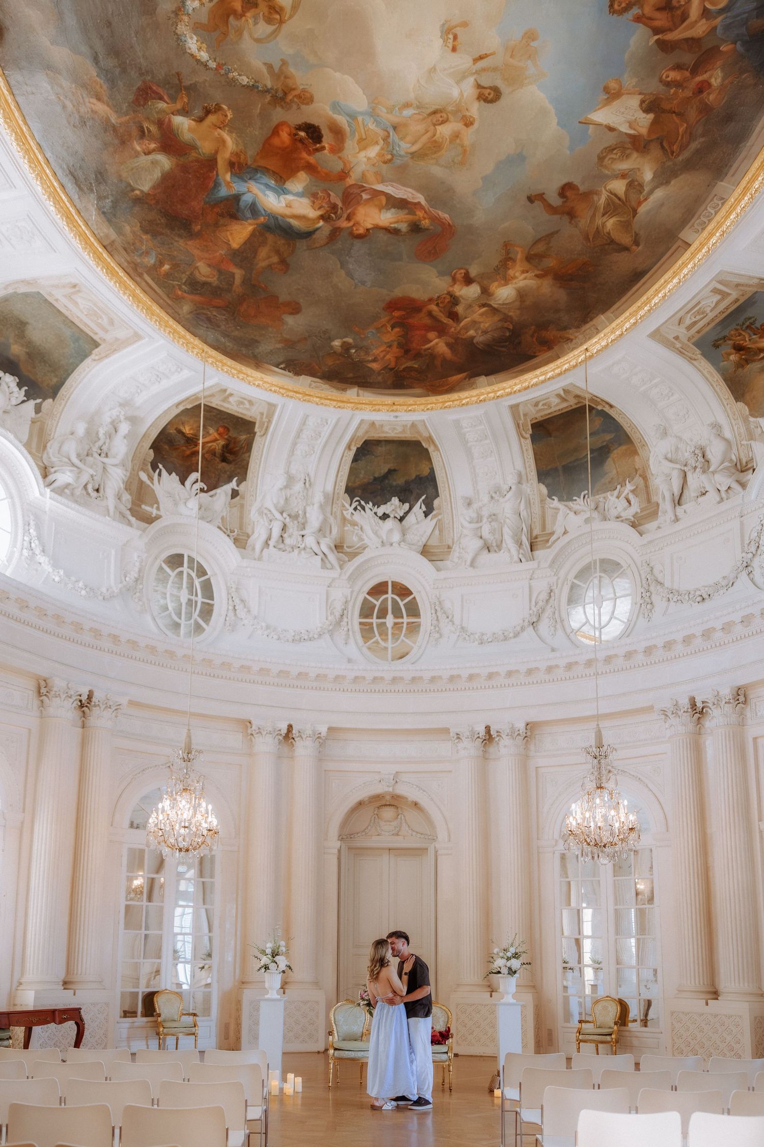 Couple embracing in ornate Baroque hall with stunning ceiling fresco, chandeliers, and white decor for an elegant wedding ceremony.