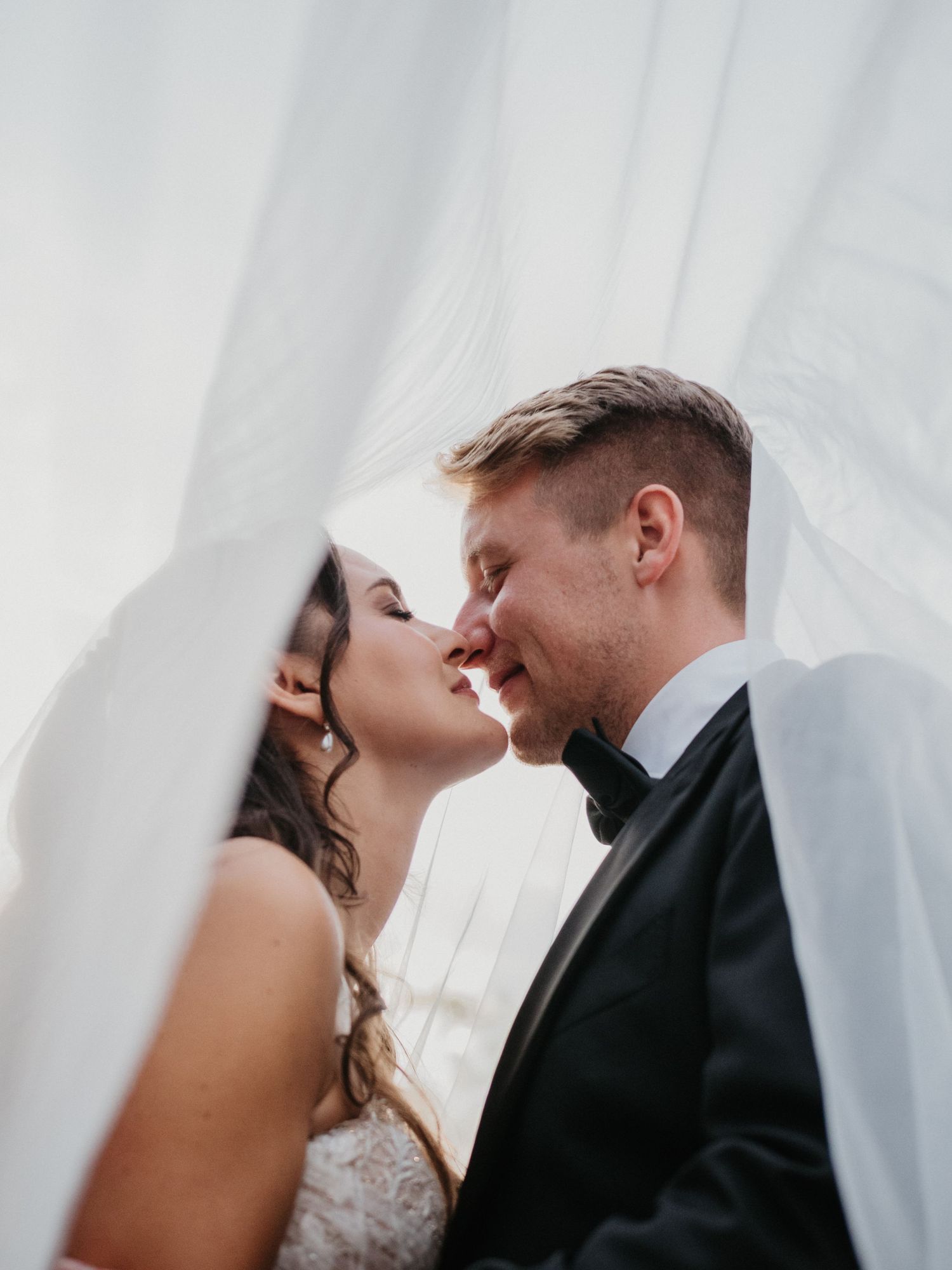 A romantic wedding photo captured through a flowing white veil as the bride and groom share an intimate moment.
