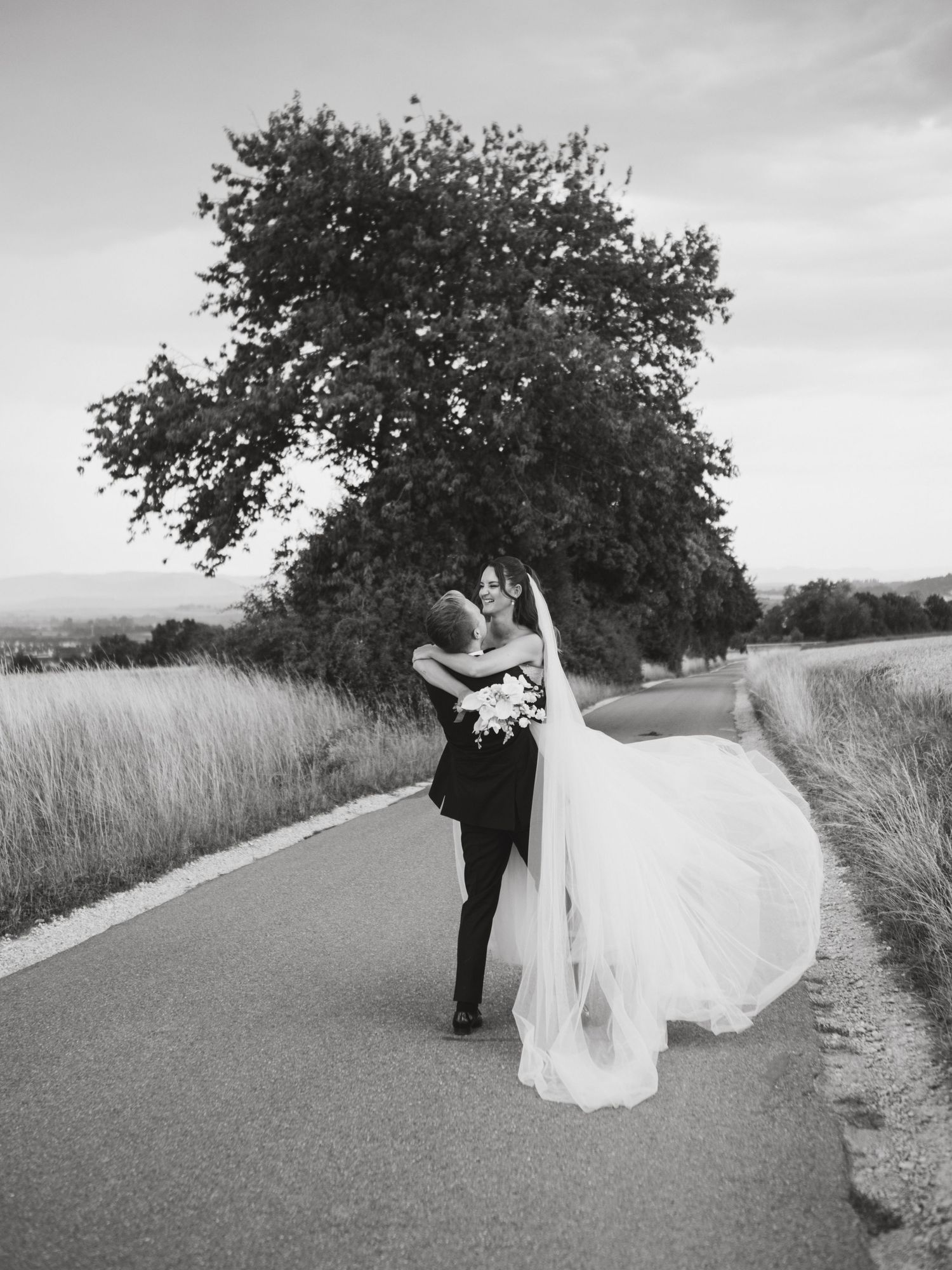 A couple in wedding attire dance together on a rural path surrounded by tall grass and trees in black and white.