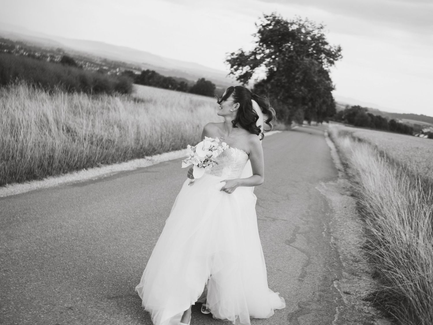 A person in a flowing white wedding dress runs along a country road in a black and white photograph.