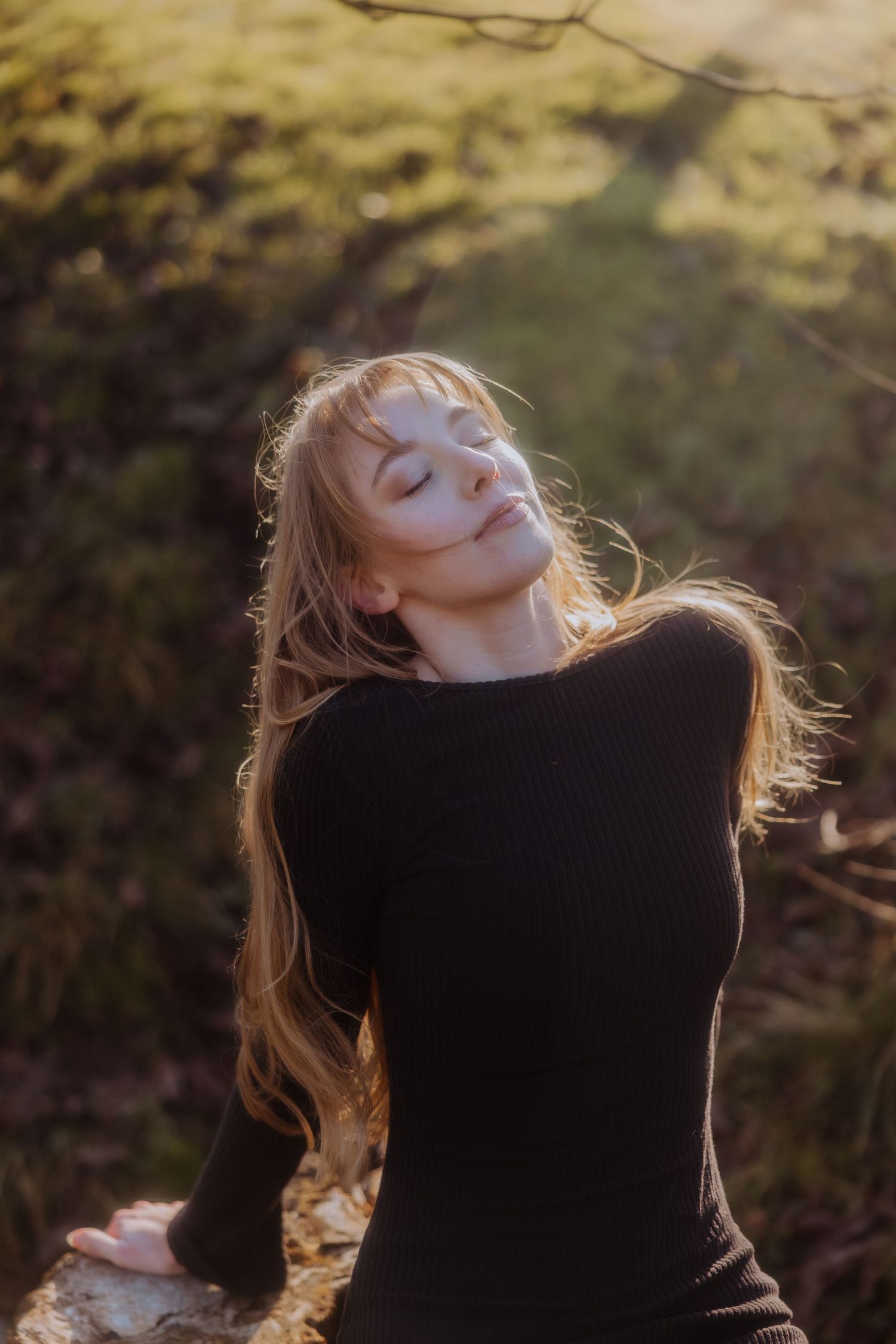 A person in a black sweater tilts their head back with hair flowing in golden sunlight through trees.