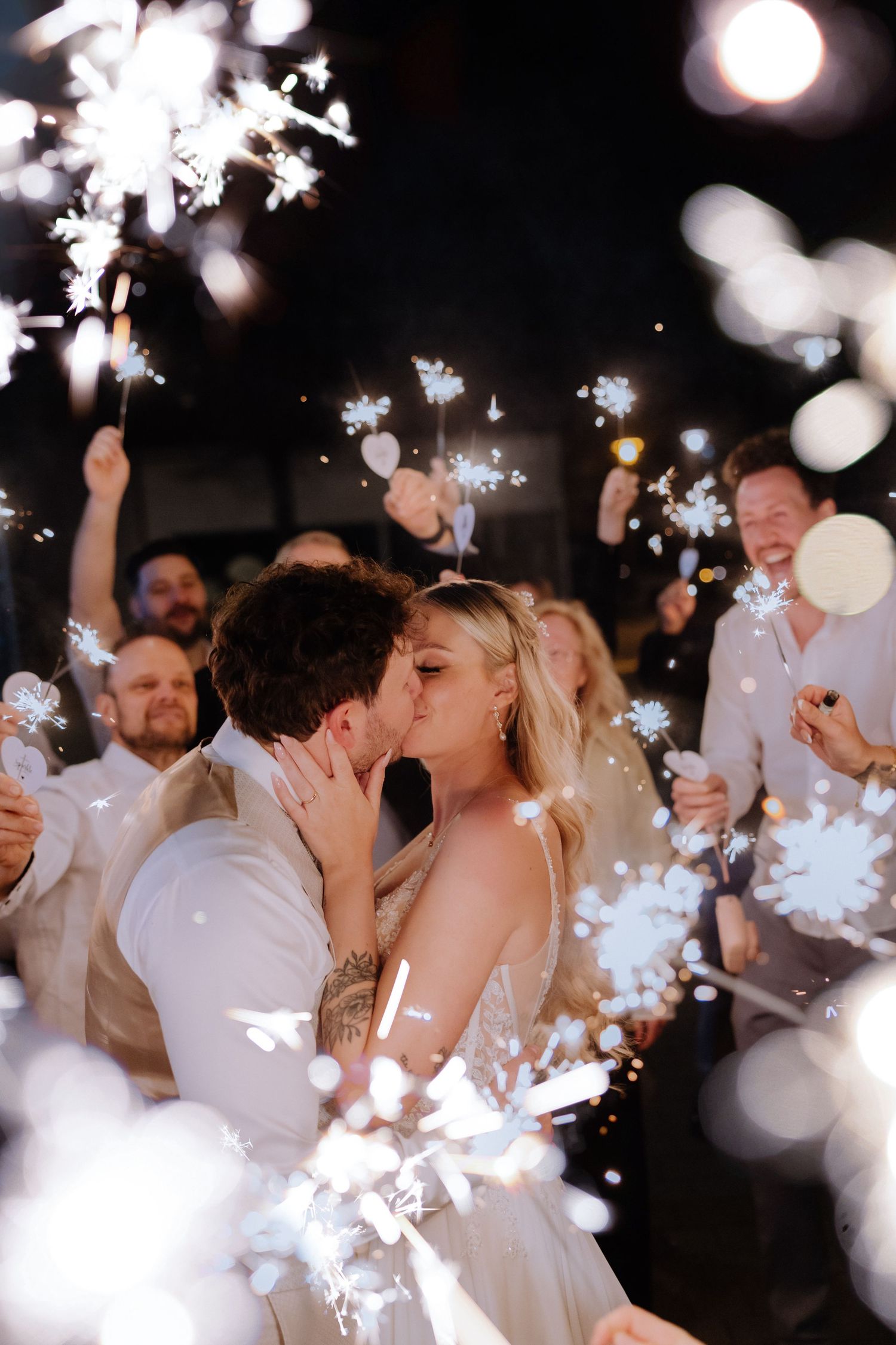 Newlyweds share a romantic kiss surrounded by guests holding sparklers during a magical wedding sendoff.