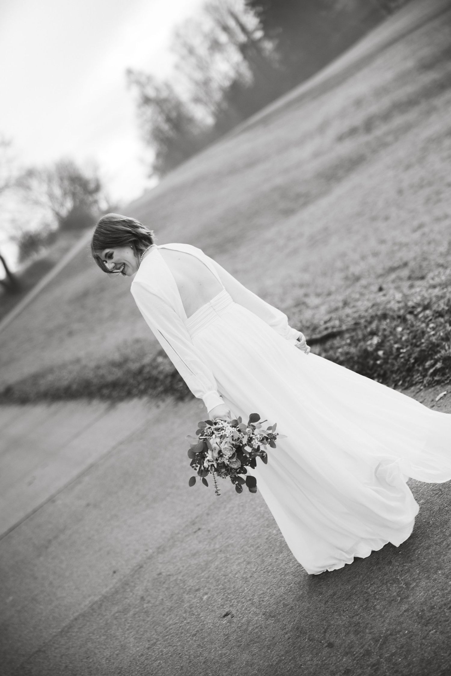 A flowing white wedding dress train billows dramatically across a grassy field in black and white photography.