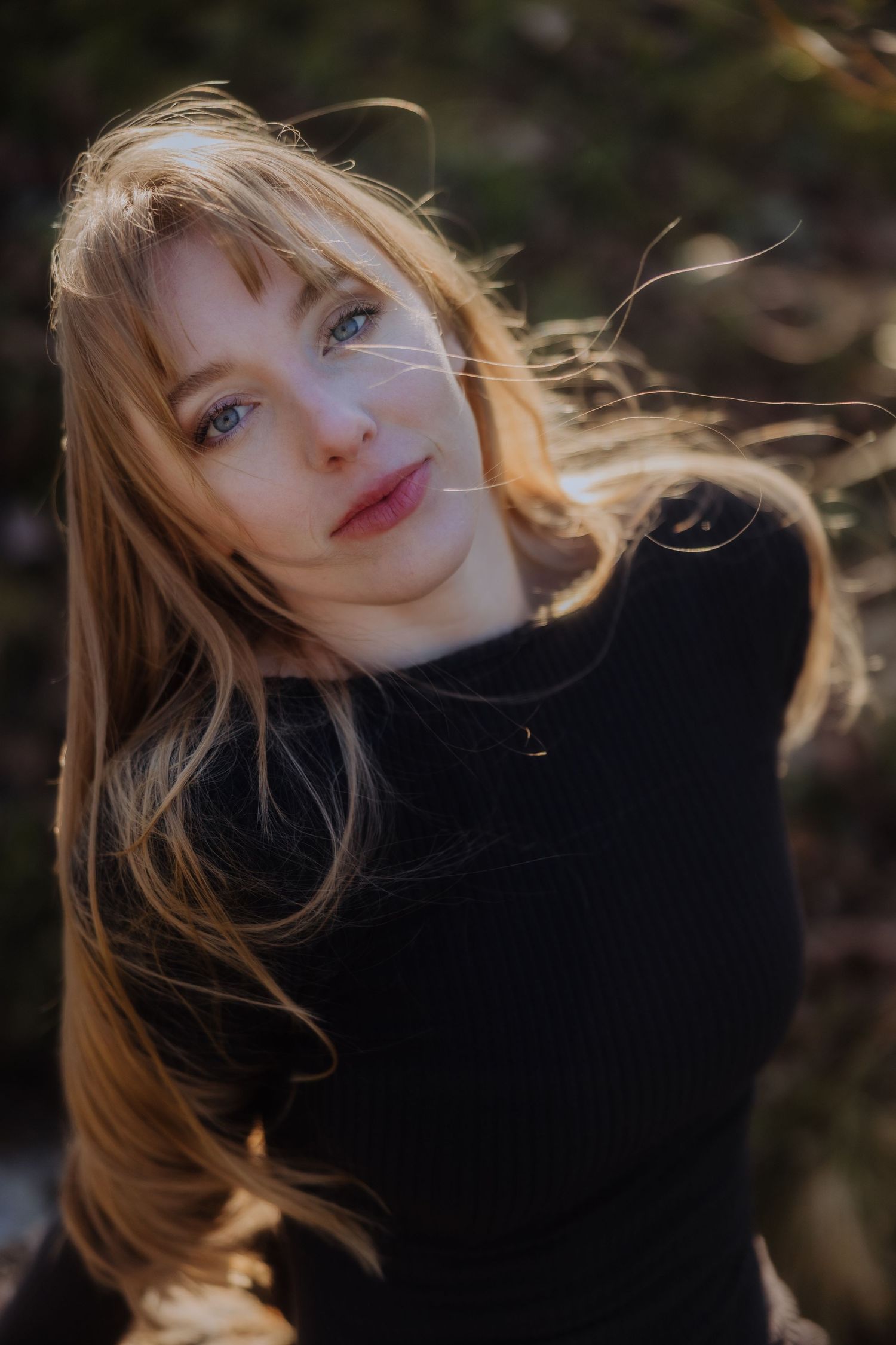 A person in a black sweater with windswept hair poses in warm natural lighting against a dark background.