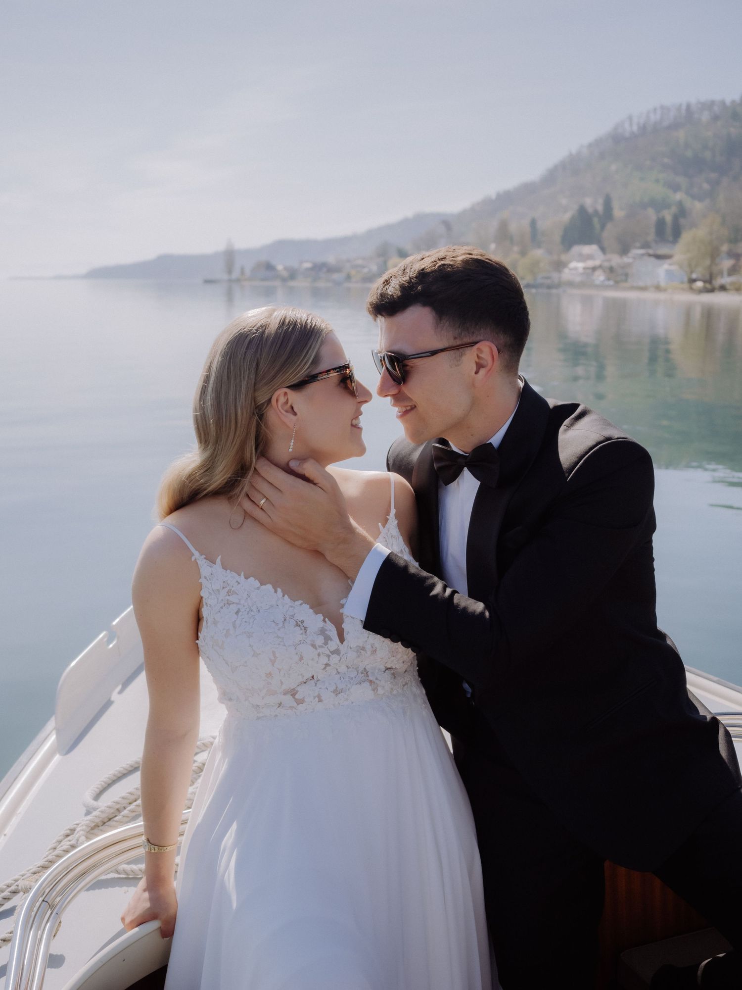 Sunlit wedding portrait by a serene lake with mountain landscape.