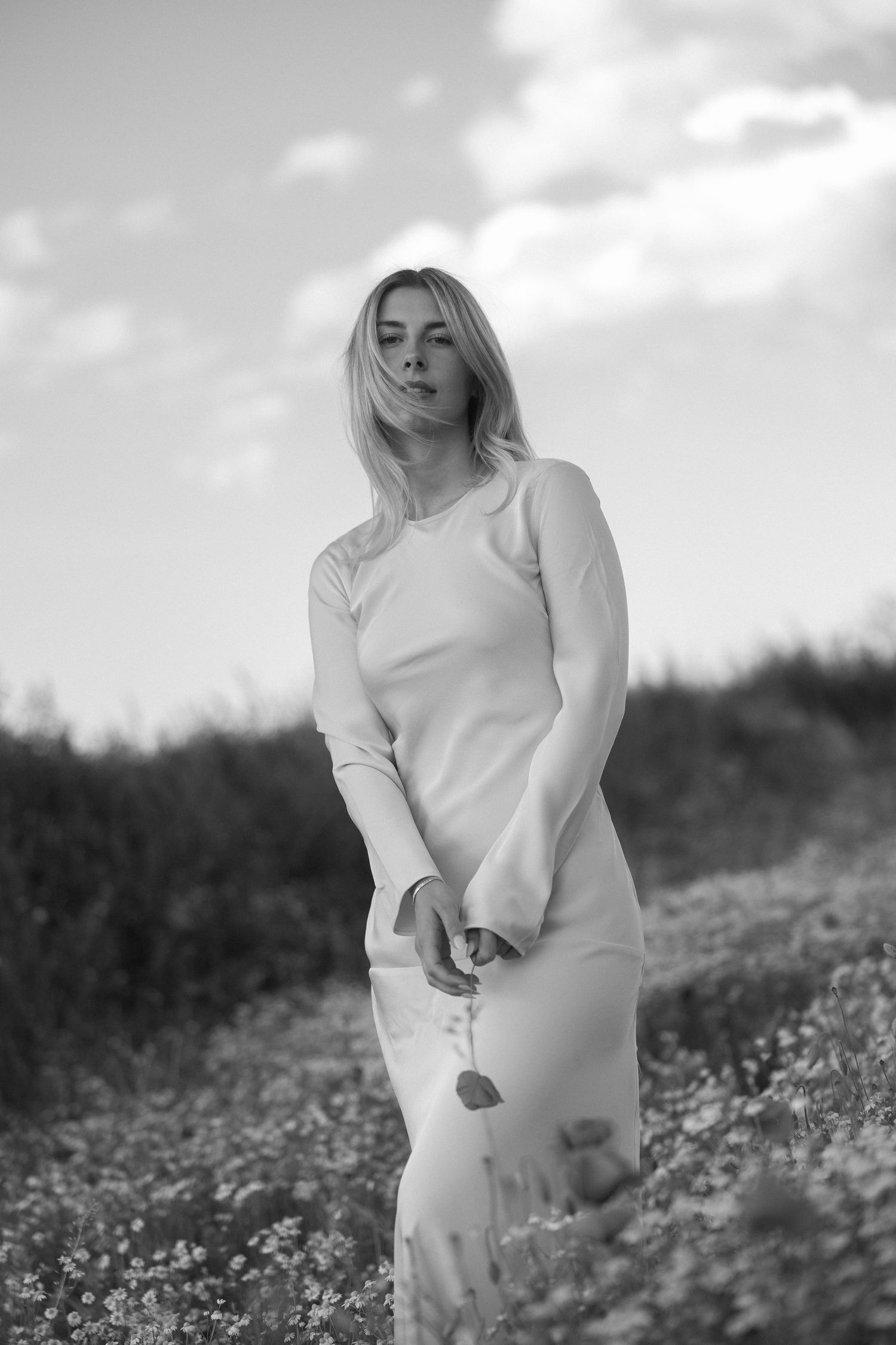 Black and white portrait of a blonde woman in a floral dress standing in a wildflower field under a cloudy sky.