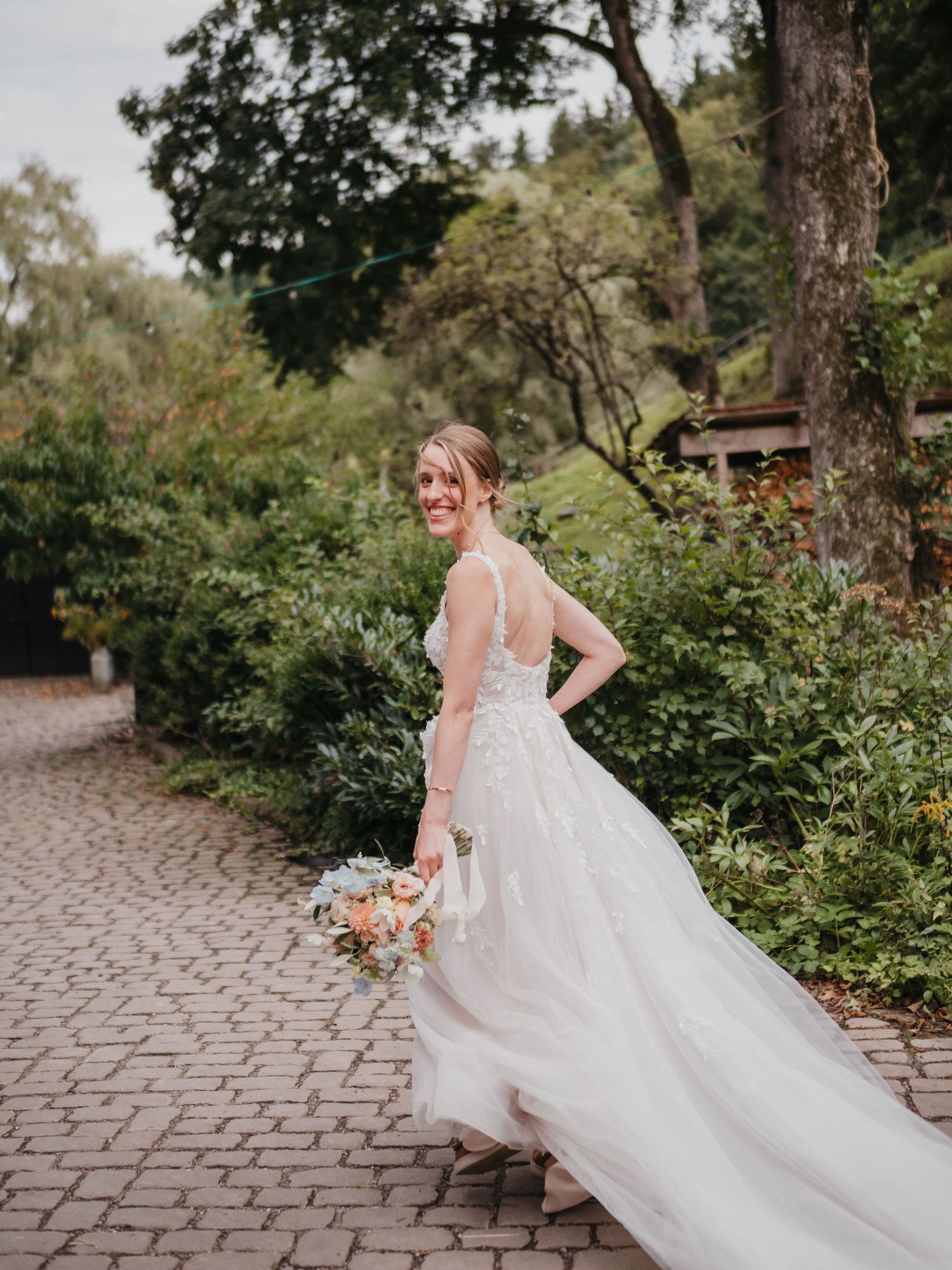 A bride in a flowing white lace gown holds a peach bouquet while standing on a stone path surrounded by lush gardens.