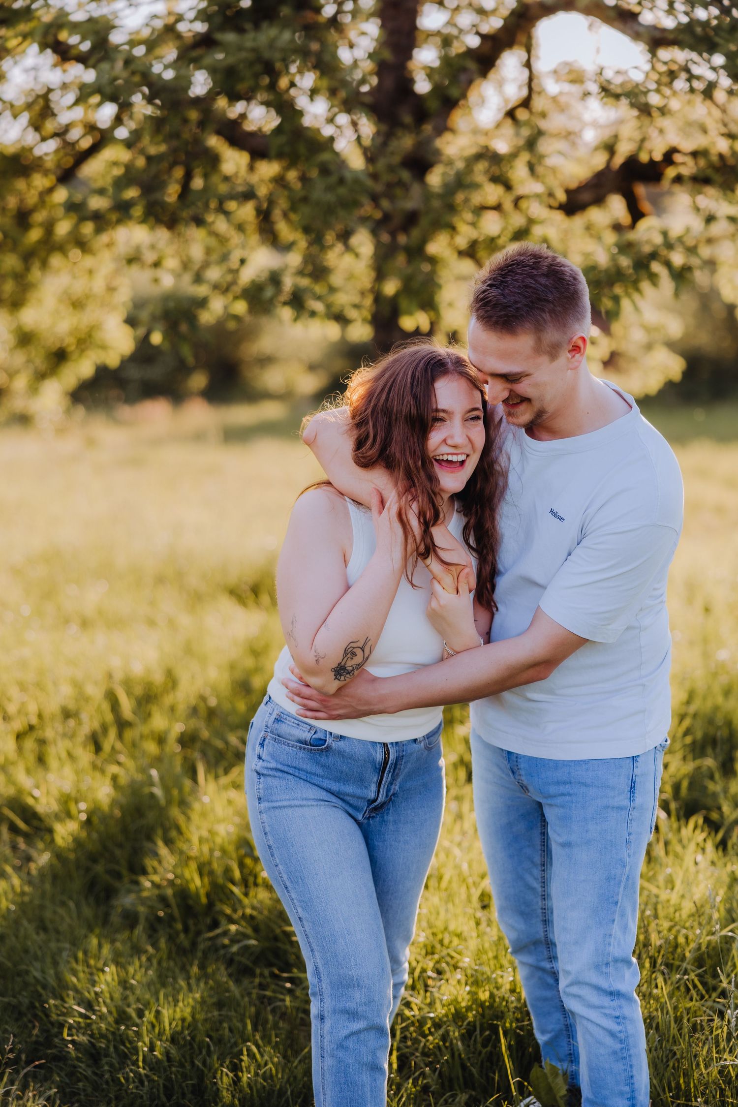 Happy couple embracing and laughing together in a sunny green field, wearing casual jeans and light tops.