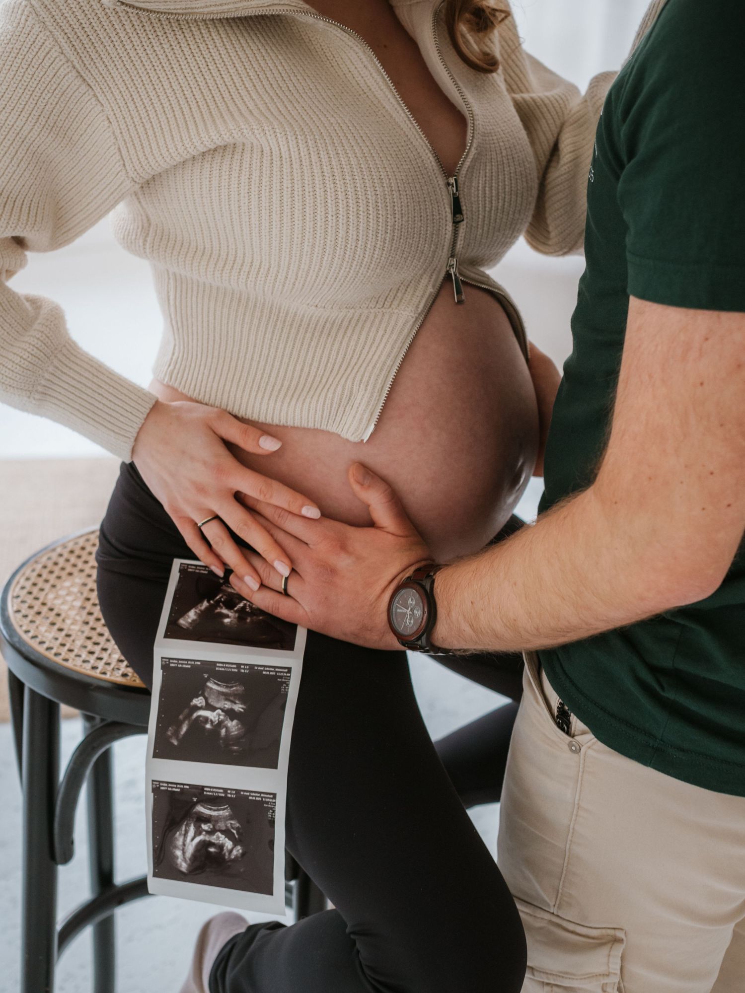 A couple holds hands over a pregnant belly while displaying ultrasound photos in a tender maternity moment.
