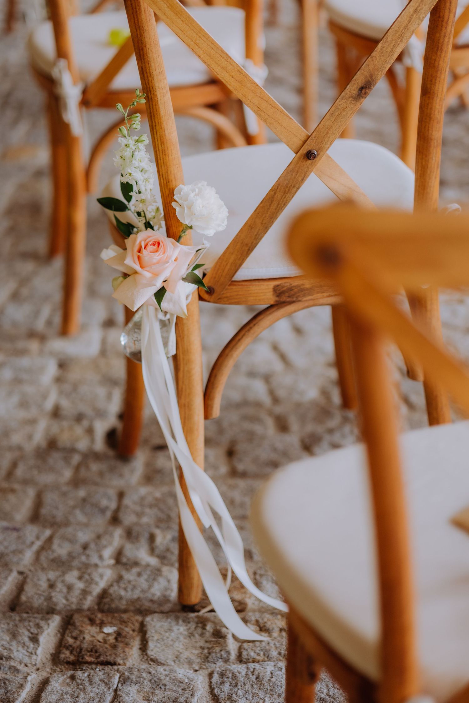 Wooden cross-back wedding chairs decorated with peach roses and white ribbon along a stone aisle.