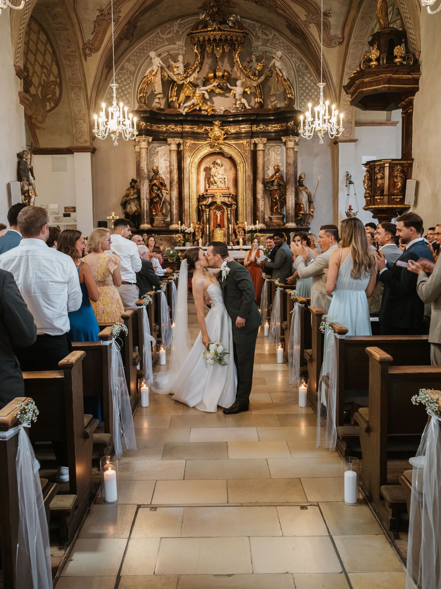 Wedding guests gather in an ornate church with chandeliers and decorative architecture during a traditional ceremony.