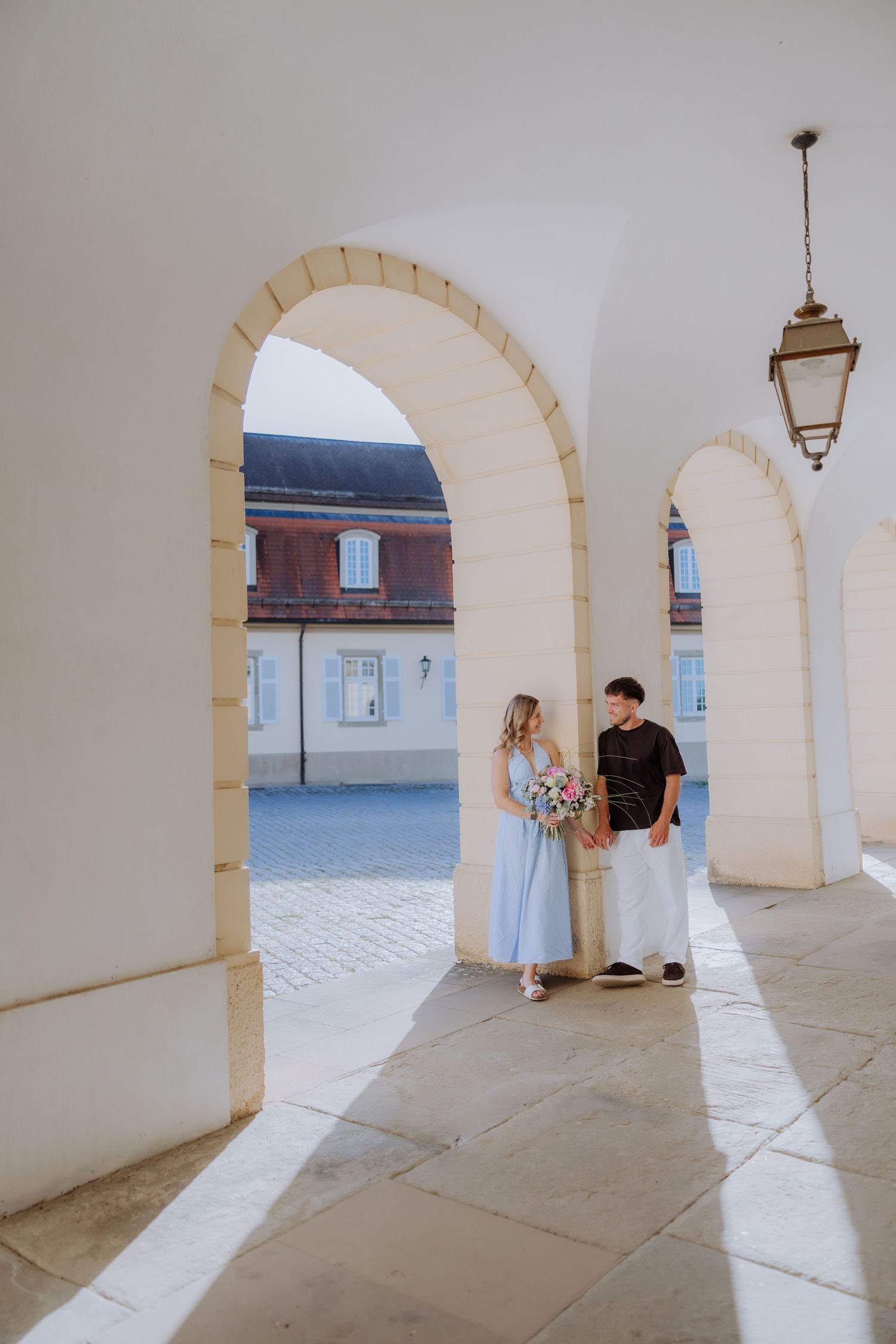 Couple holding flowers under white arched colonnade of elegant European building on sunny day.