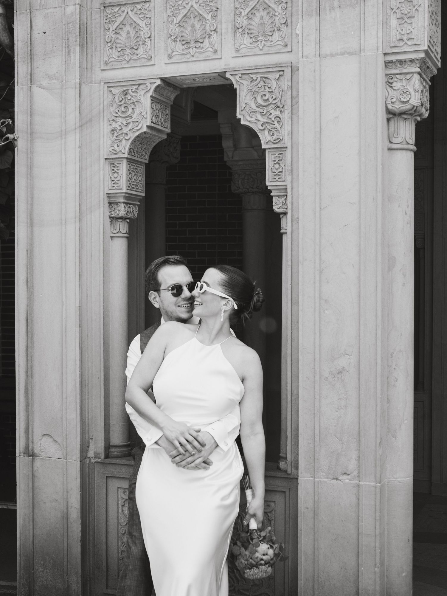 A romantic black and white photo series showing a couple in white attire embracing in front of ornate architectural details.