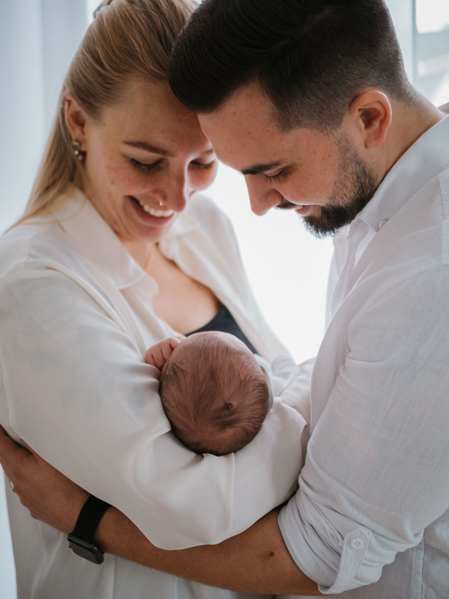 A couple in white clothing sharing tender moments with their newborn baby in a bright room.