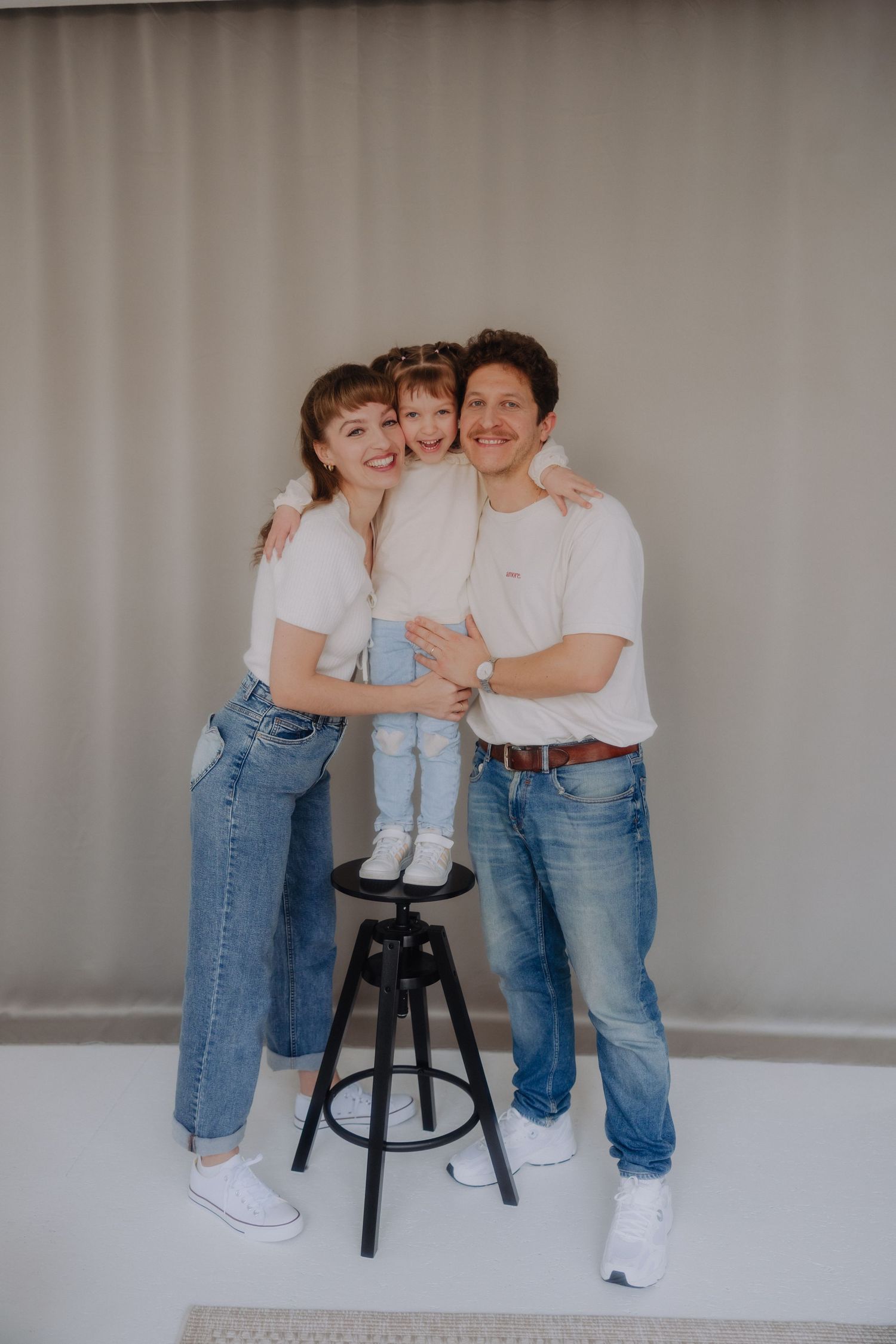 Family wearing matching white tops and blue jeans pose together against grey background with child standing on stool.