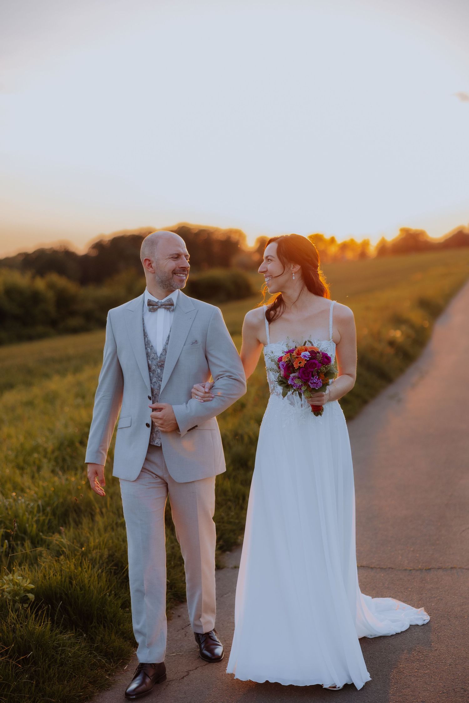 Bride and groom walking together at sunset on a path, she holds a colorful bouquet in a white gown, he wears a gray suit.