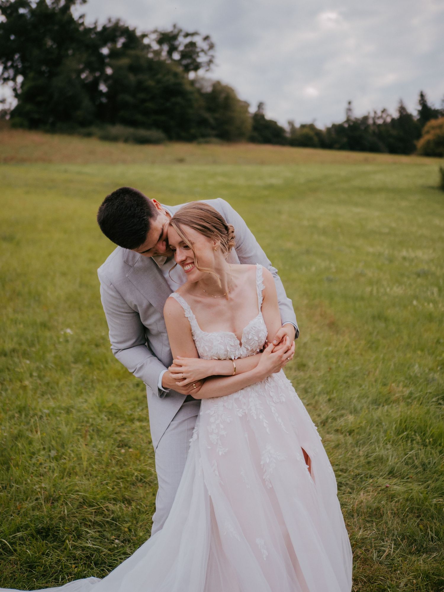 Romantic wedding couple embracing in a sunlit meadow wearing white and grey attire.