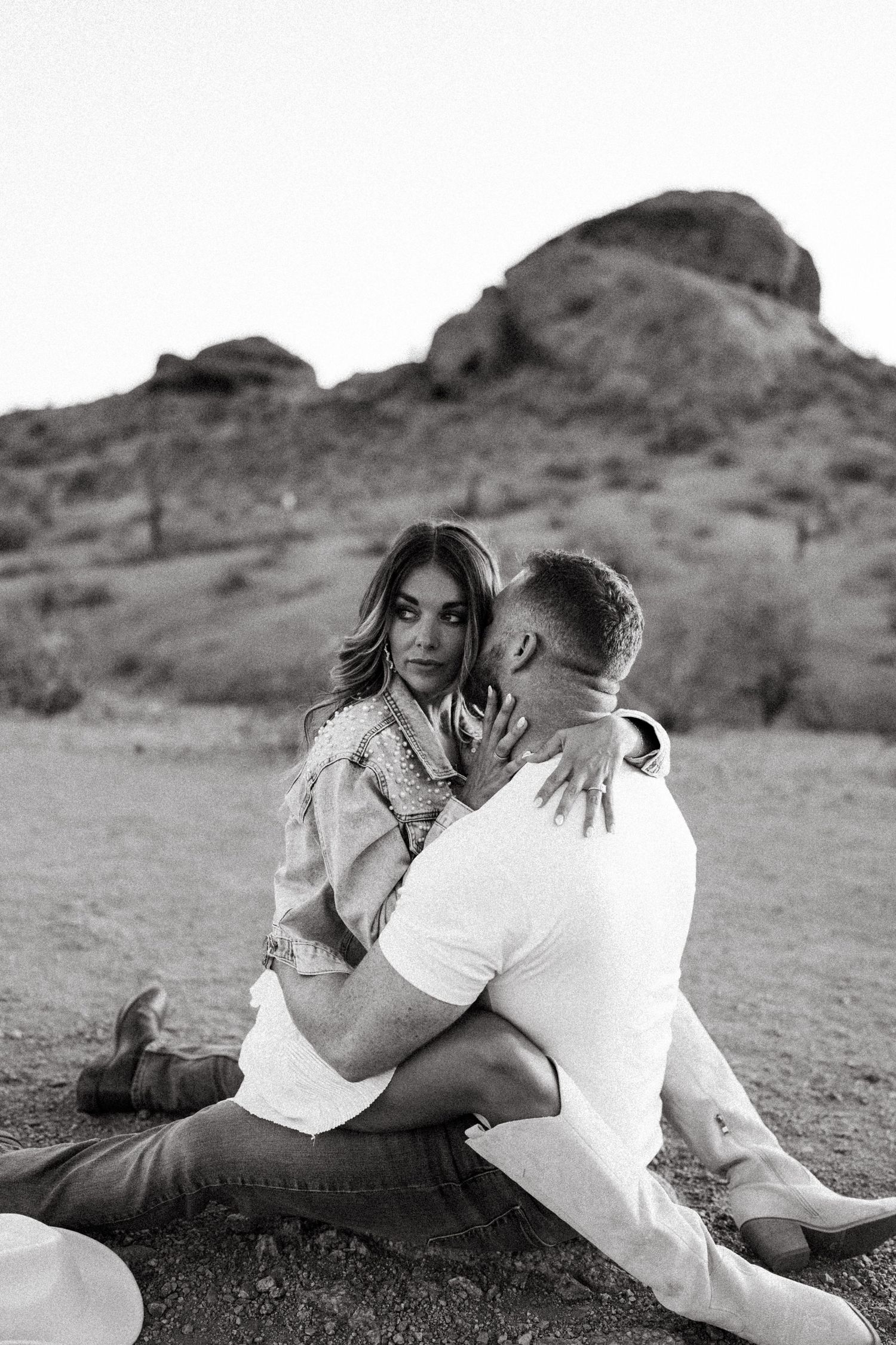 A black and white portrait of an intimate couple embracing against a desert mountain landscape.