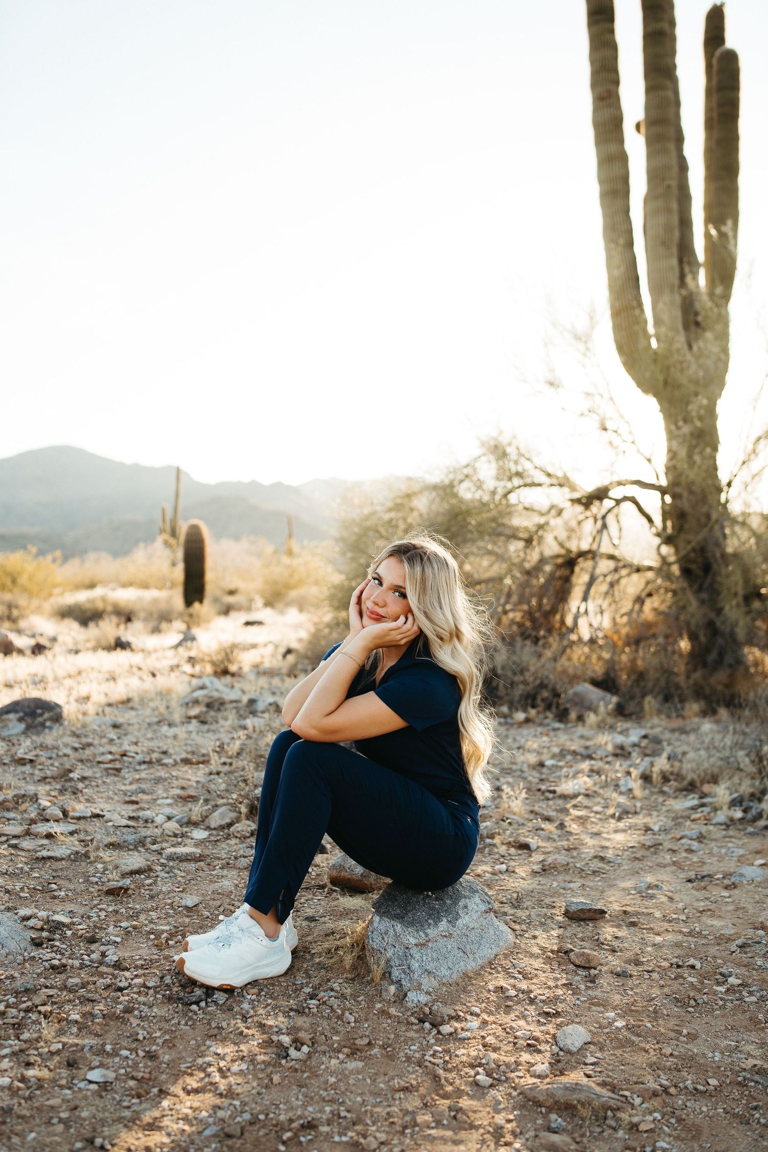 Person in casual attire sits thoughtfully on rock beneath tall saguaro cactus in desert setting.