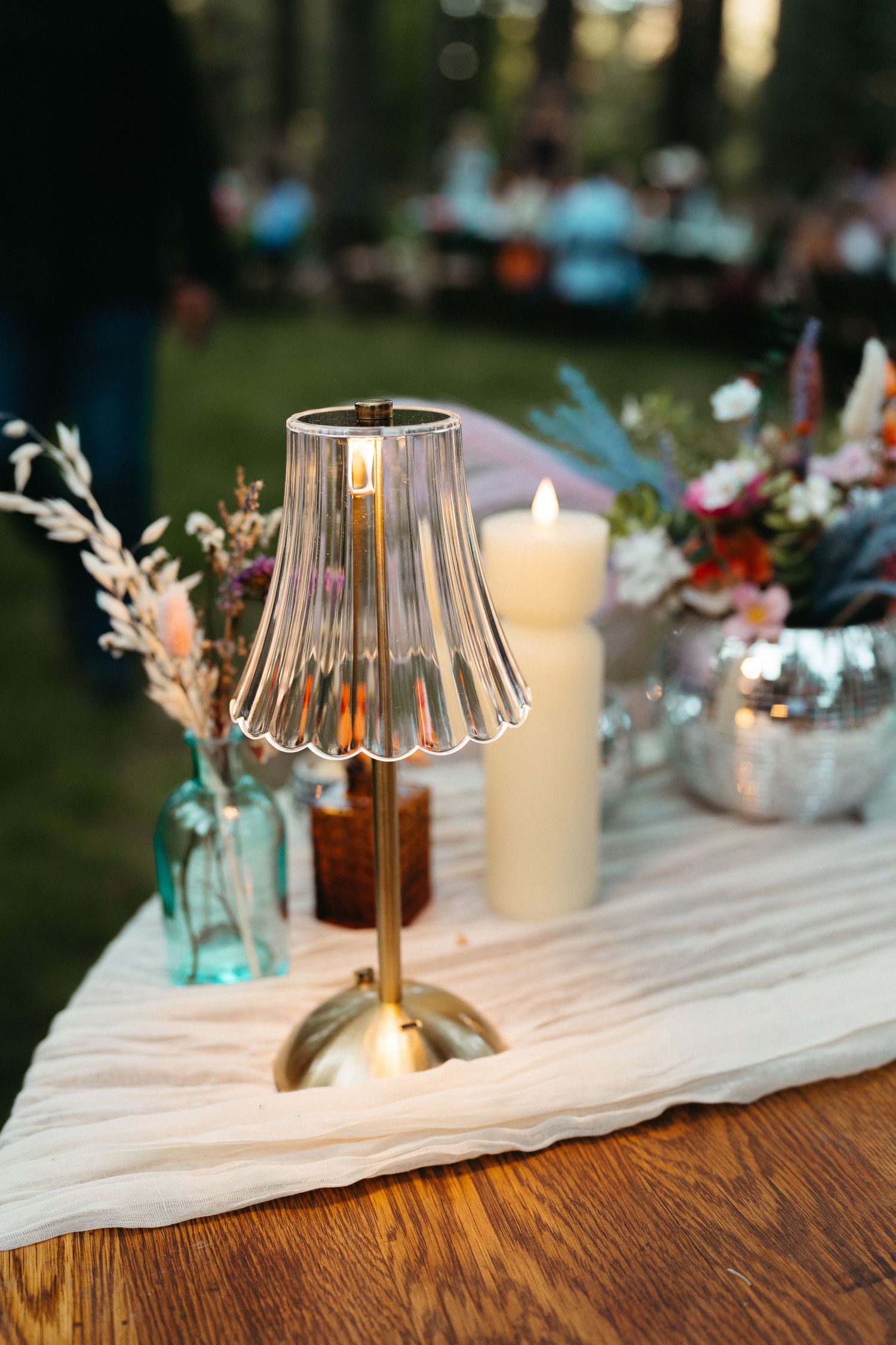 A vintage-style glass lamp with brass base sits on a wooden table with floral decorations and candles at an outdoor event.