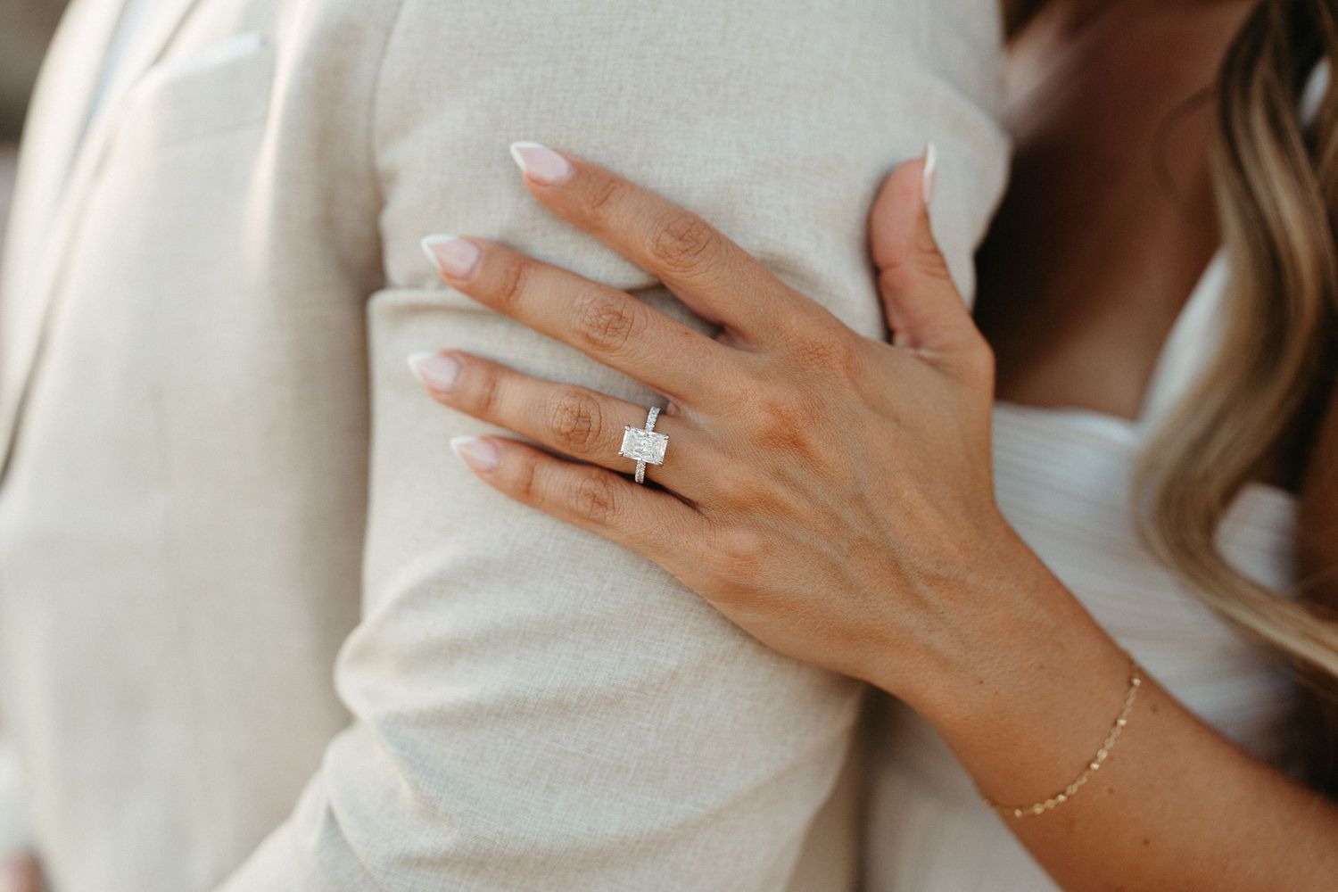 A delicate hand wearing a sparkling engagement ring and dainty bracelet rests against a soft white sweater.