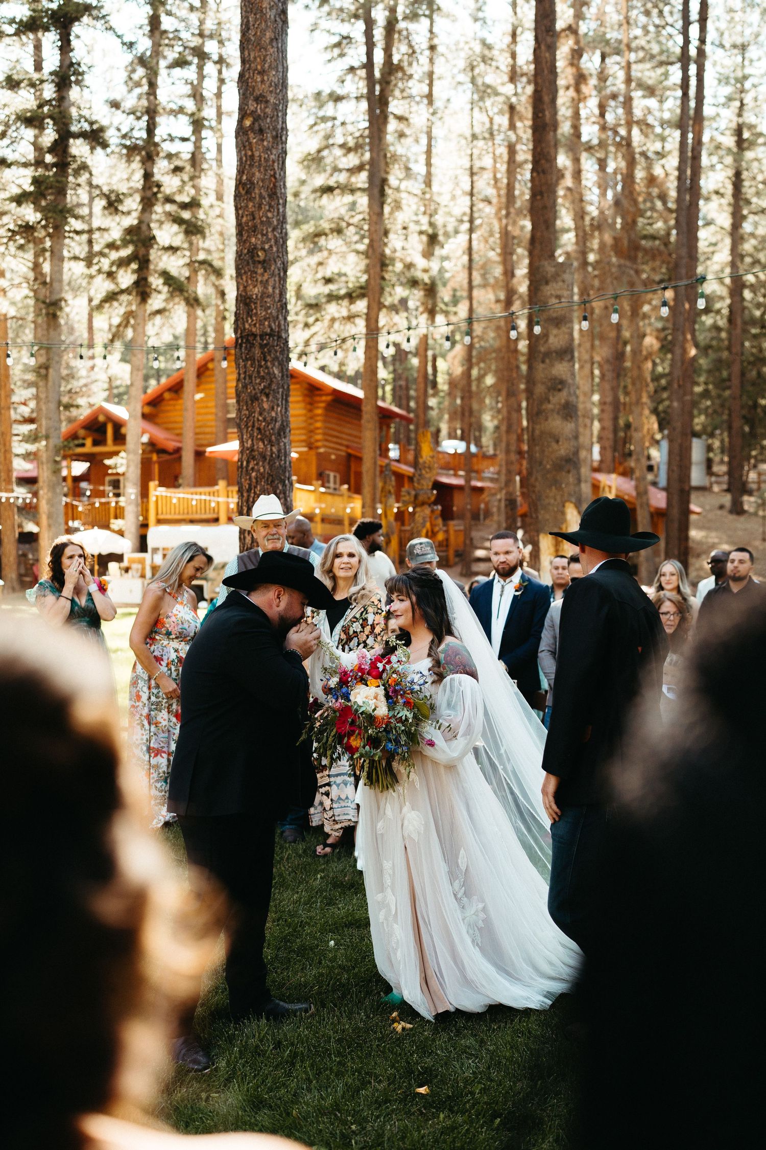 A romantic wedding ceremony takes place in a forest setting with wooden cabins visible in the background.