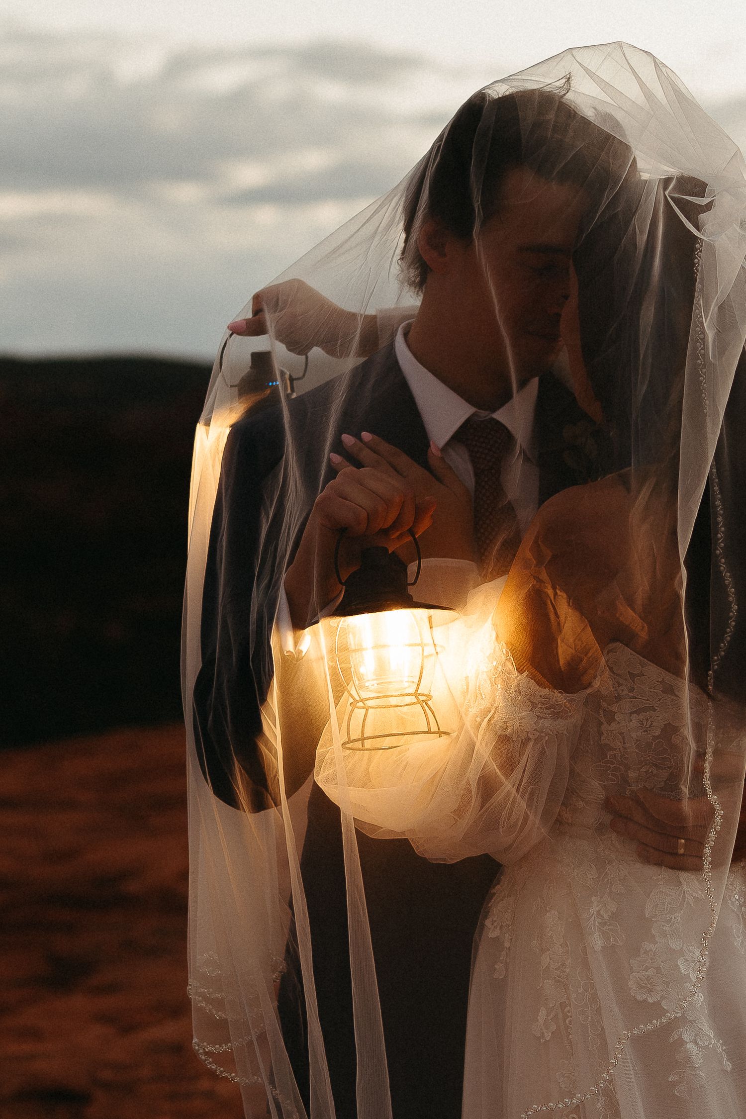 Romantic wedding couple embraces under a veil at sunset with glowing light creating a magical silhouette effect.