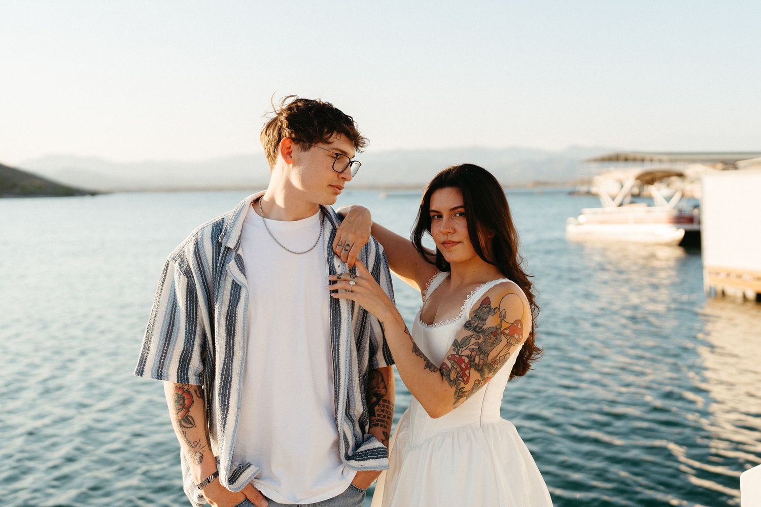 Romantic couple in casual attire embracing by tranquil waterfront during golden hour with boats in background.