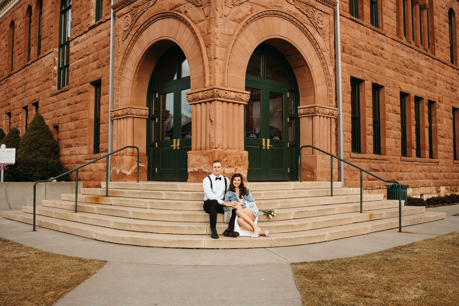 A couple sits on the steps of a historic red brick building with arched doorways.