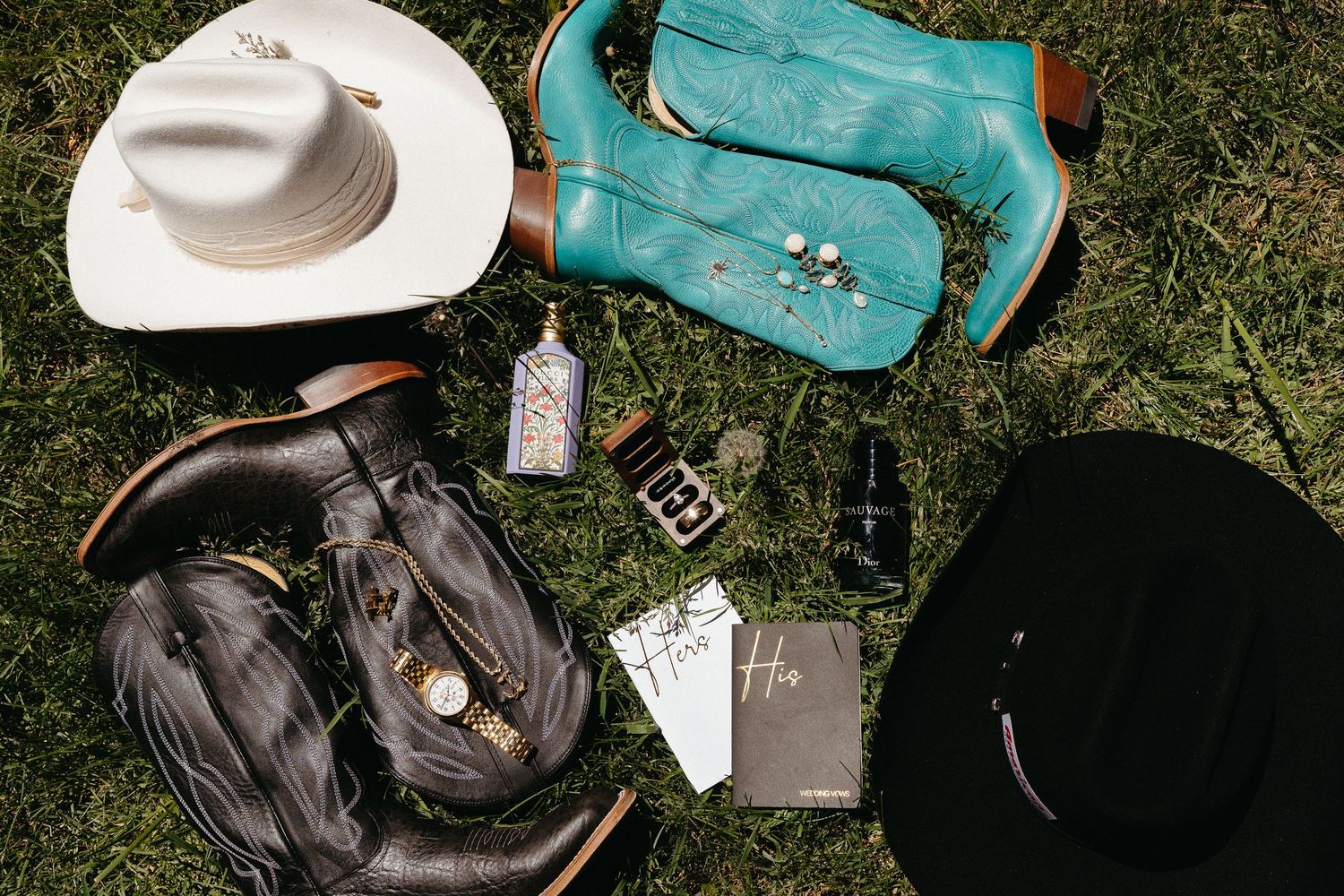 Cowboy hats, boots, and western accessories laid out on grass, including a turquoise leather bag and playing cards.