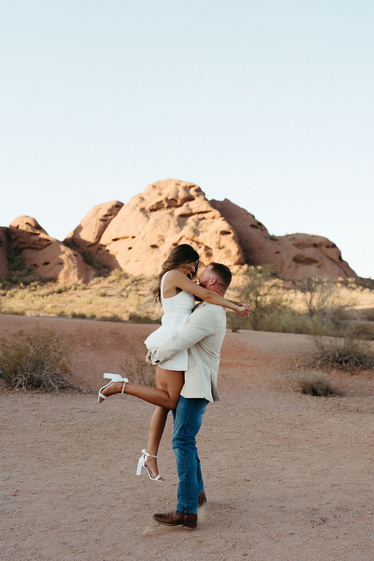 Two people embrace passionately in a romantic desert setting at sunset with red rock formations in the background.