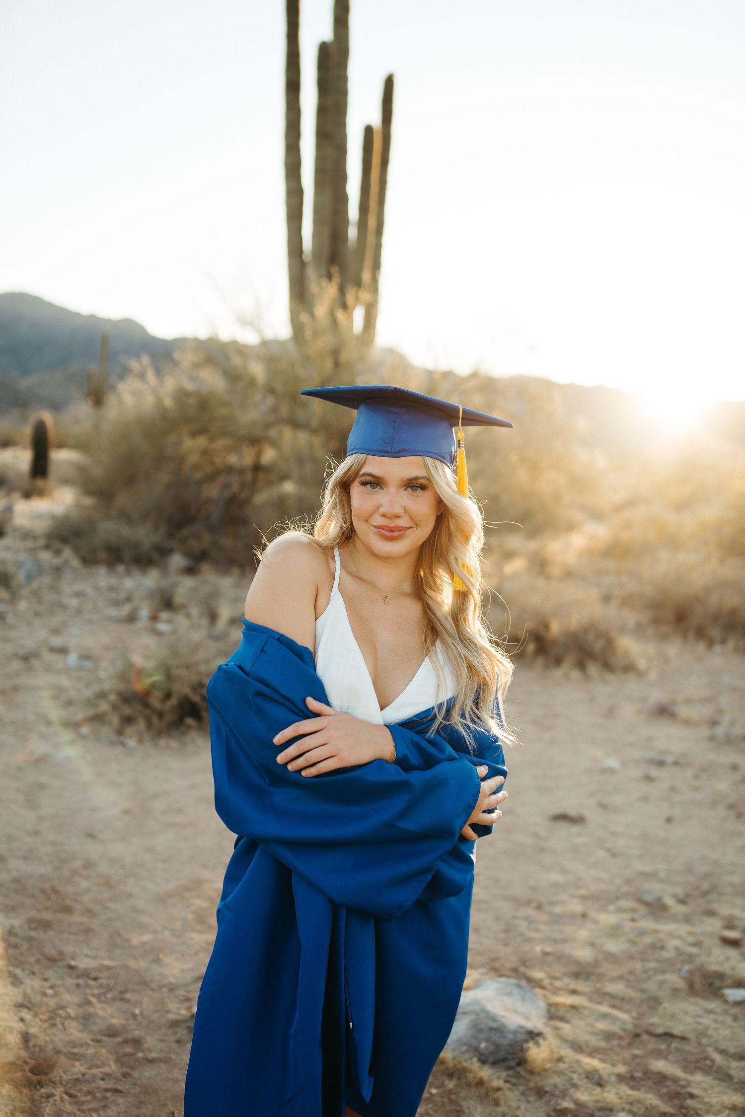Graduate in blue cap and gown poses in front of saguaro cactus at sunset.