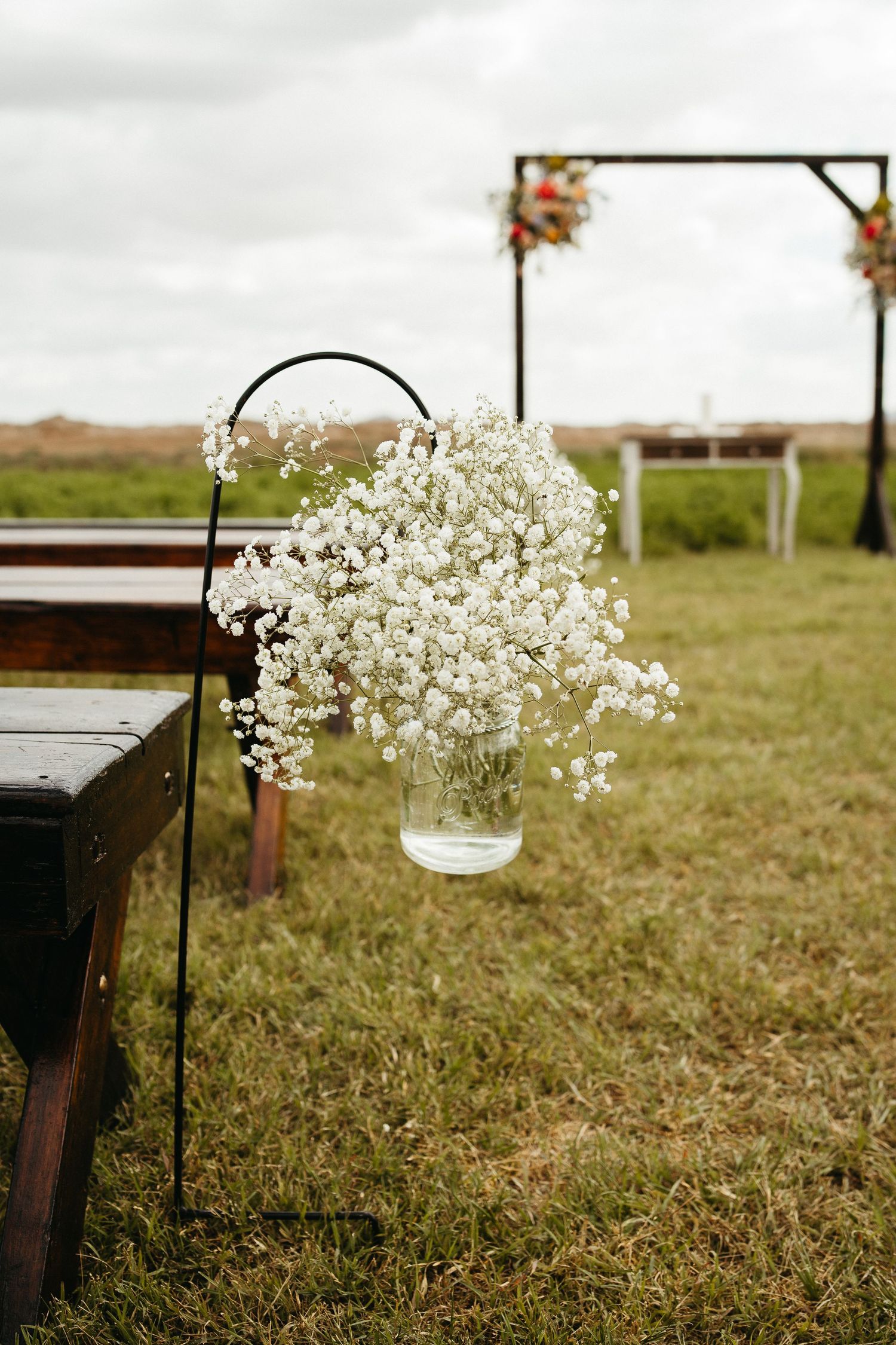 White baby's breath flowers in mason jar hang from shepherd's hook along rustic outdoor wedding aisle.
