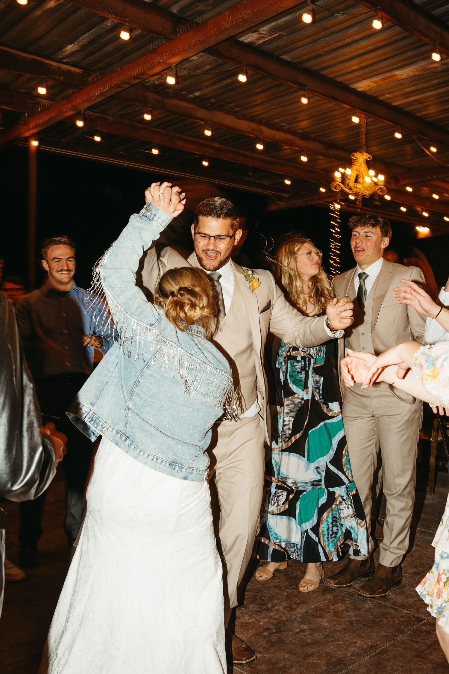 Wedding guests dancing and celebrating under wooden beams with string lights at an indoor reception.