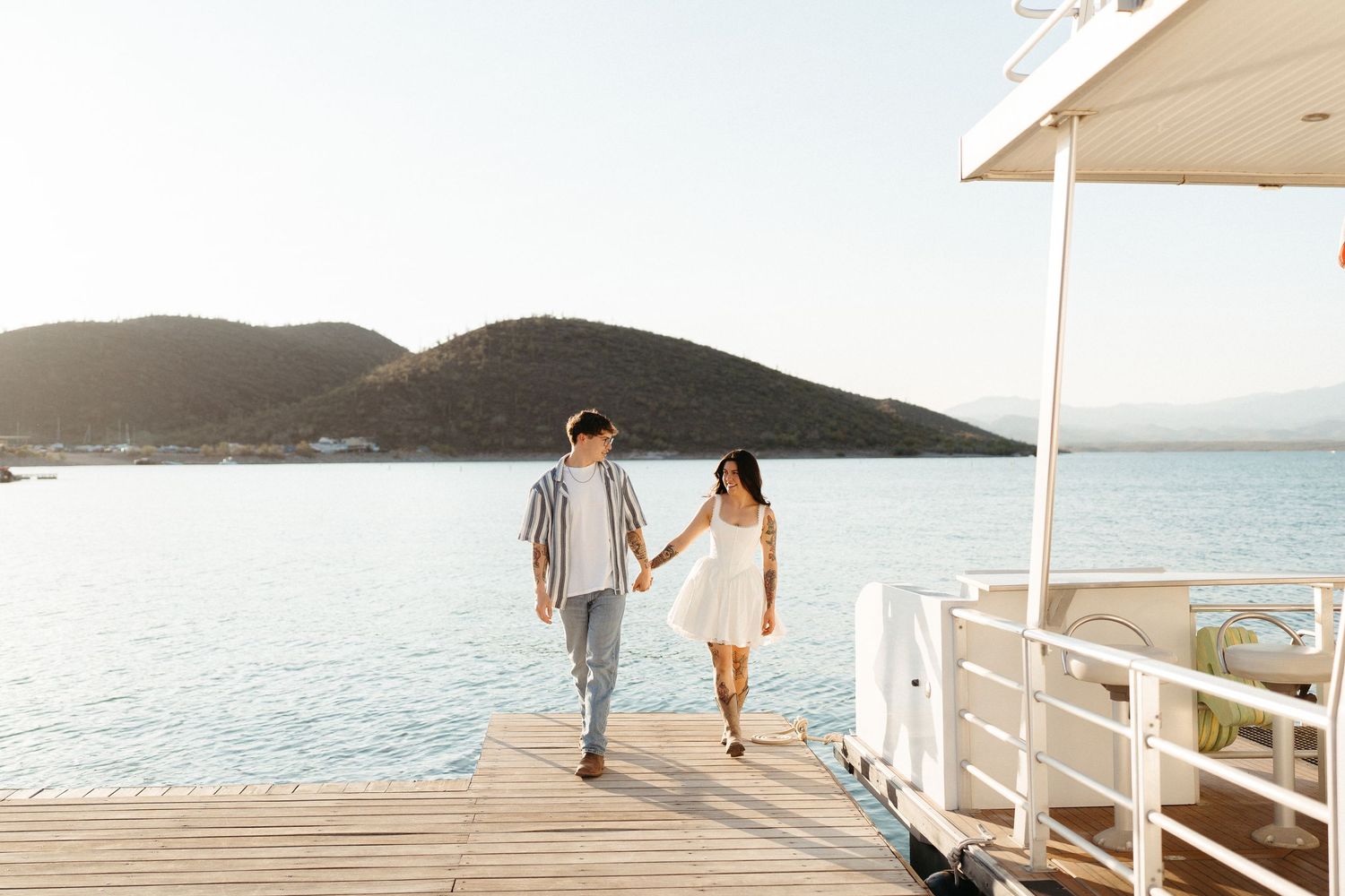 A couple strolls along a scenic white boardwalk overlooking a calm ocean and mountain landscape at sunset.