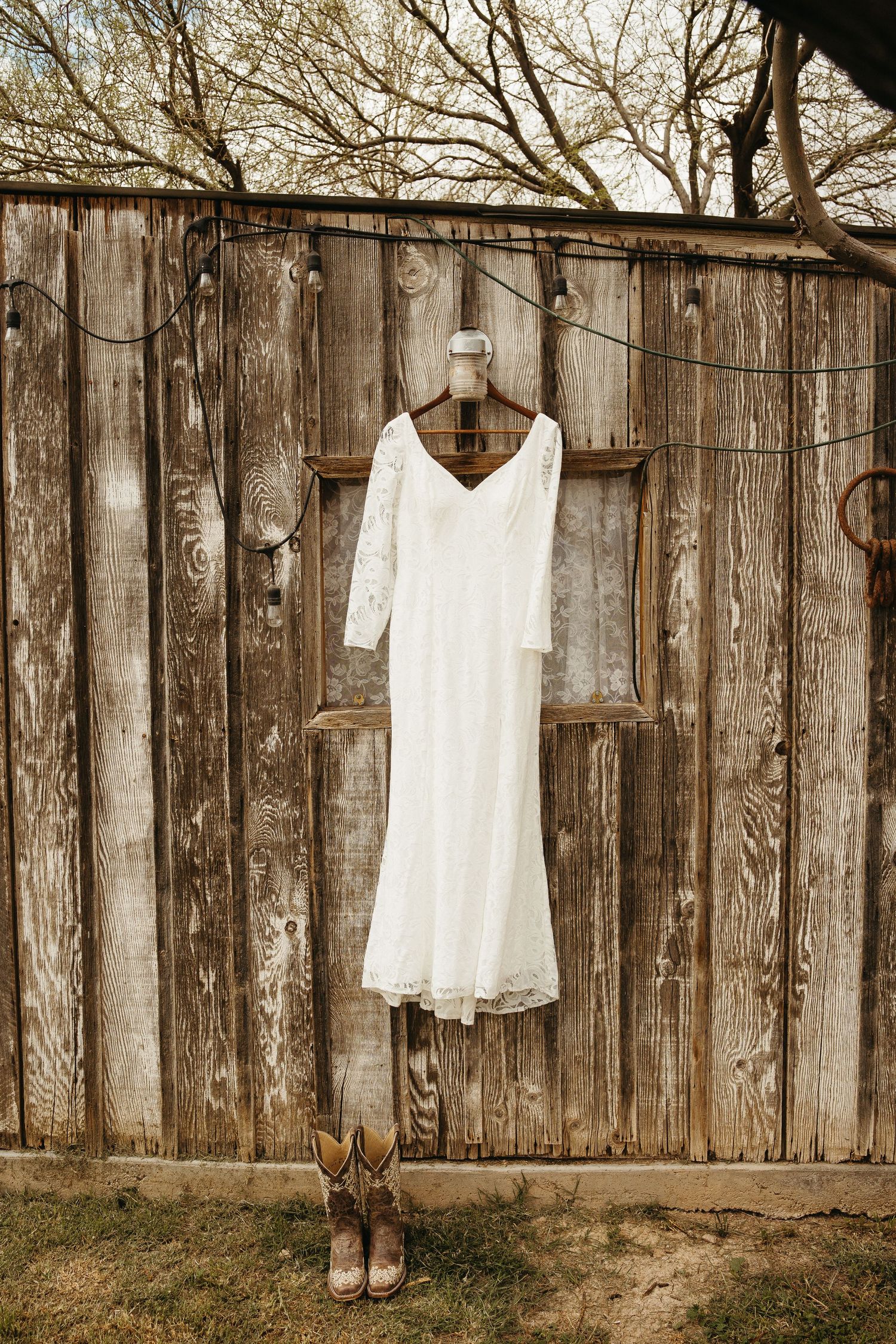 White lace wedding dress hanging on rustic wooden fence with embroidered cowboy boots below.
