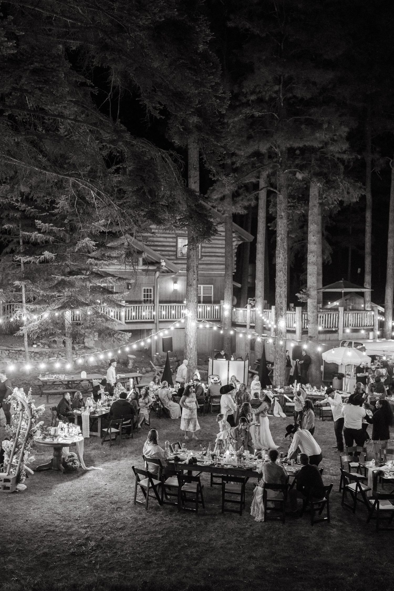 Black and white outdoor wedding reception with string lights and tables arranged under tall trees in a forest setting.