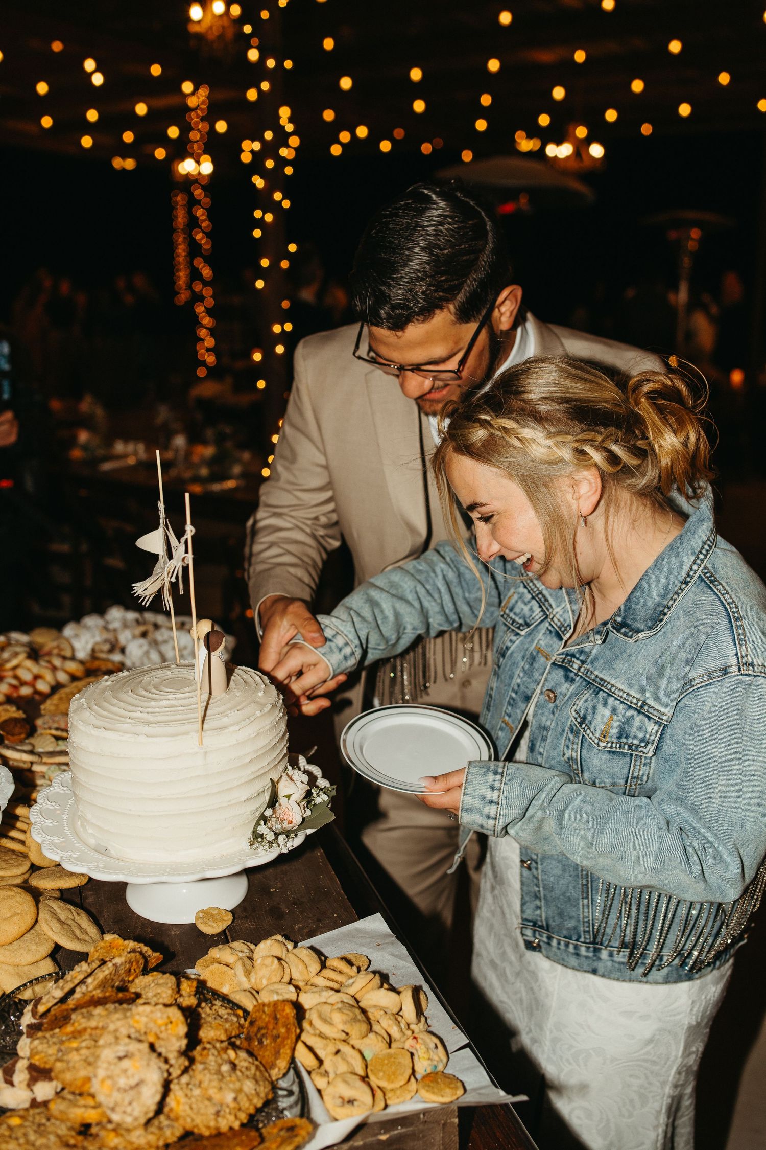 A couple cuts their white wedding cake together at a dessert table with cookies.