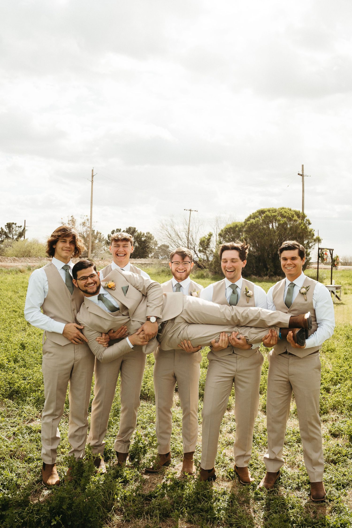Groomsmen in beige suits playfully carry one of their group members during outdoor wedding photos.