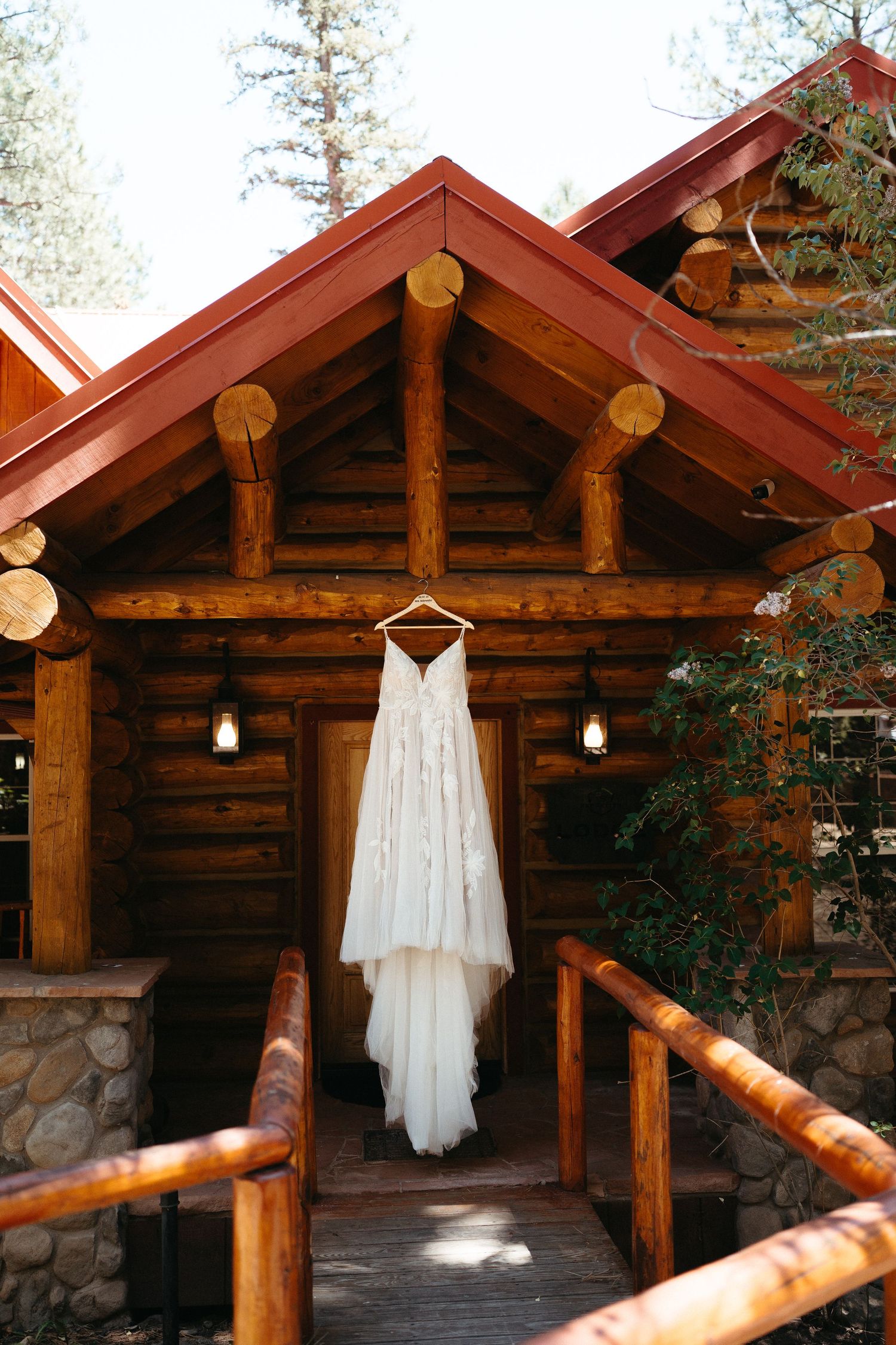 A white wedding dress hangs on display outside a rustic wooden log cabin with stone foundation and red roof.