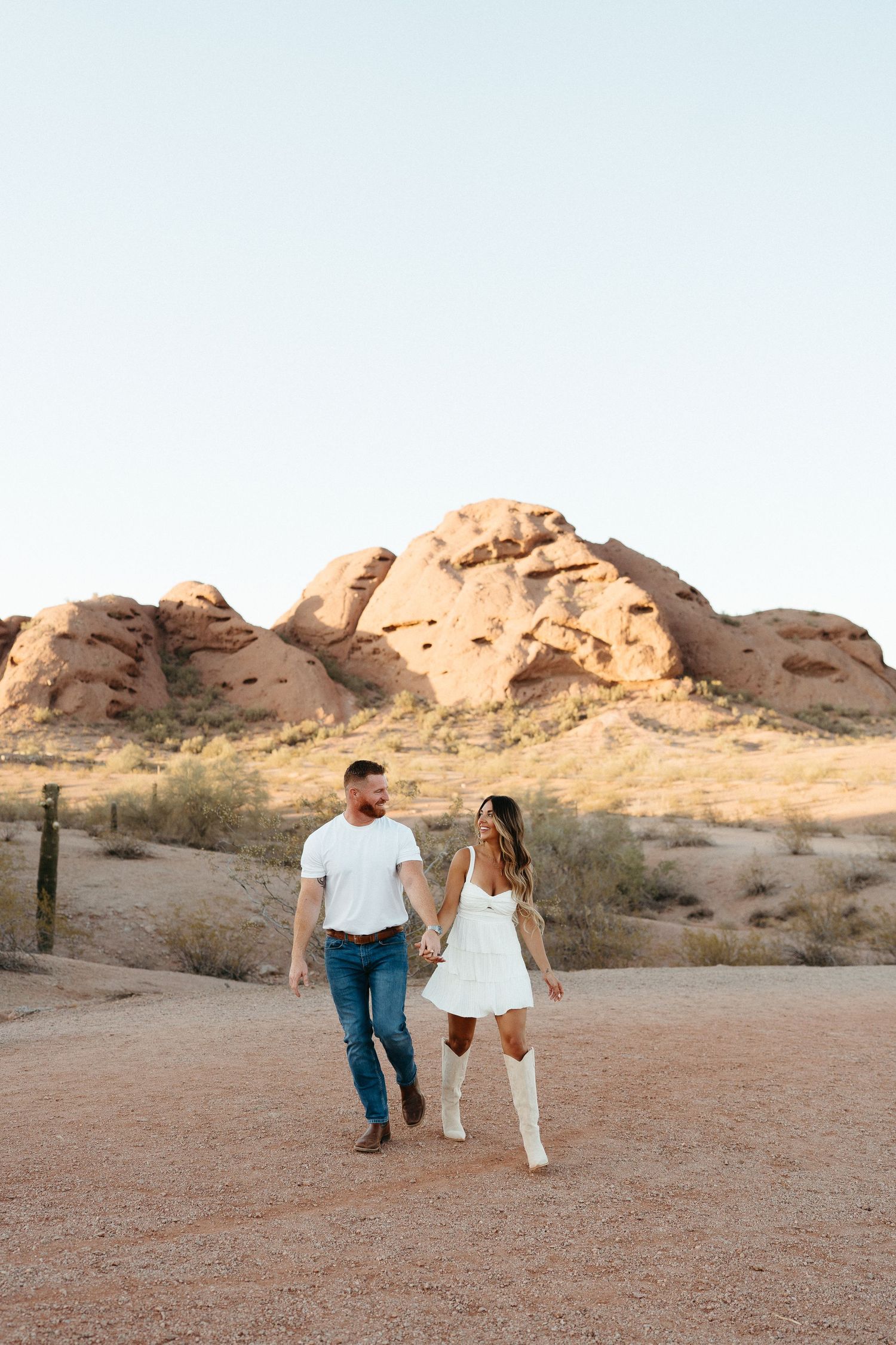 Couple walks hand in hand through desert landscape with red rock formations during sunset engagement session.