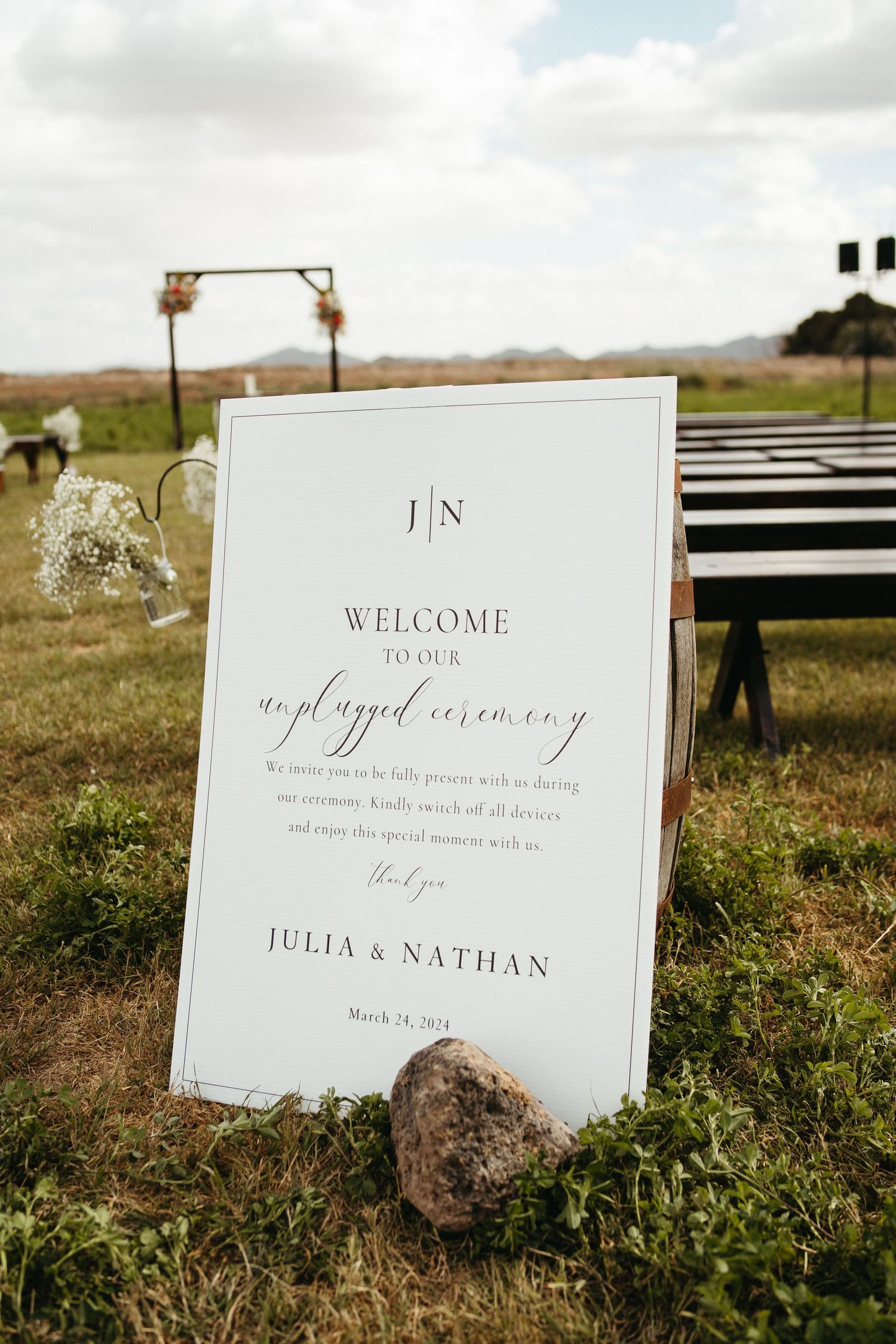Elegant white welcome sign for Julia and Nathan's wedding displayed on grass with wooden arch in background.